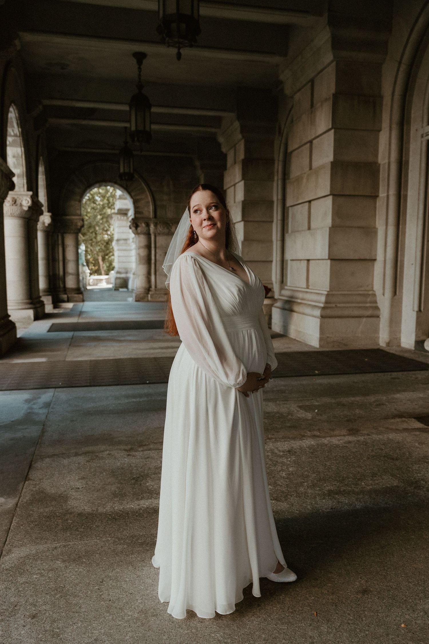 A bride in a flowing white dress poses in an elegant stone colonnade with arched architecture.
