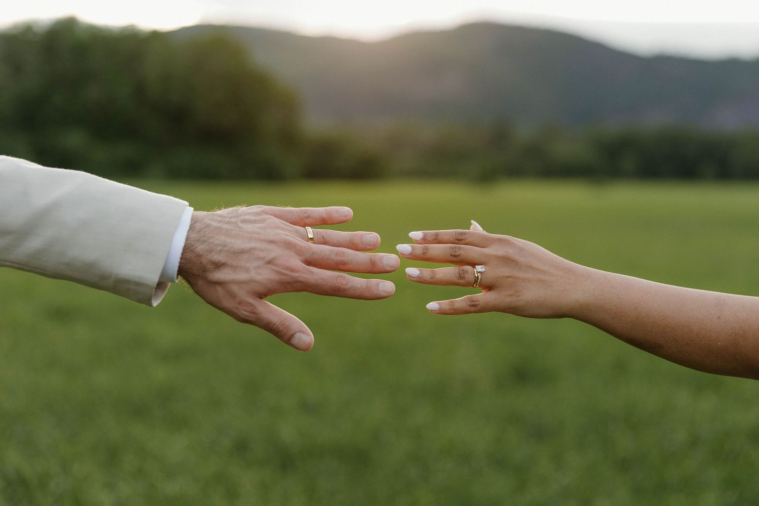 Two hands with wedding rings reaching toward each other over a green field with mountains at sunset.