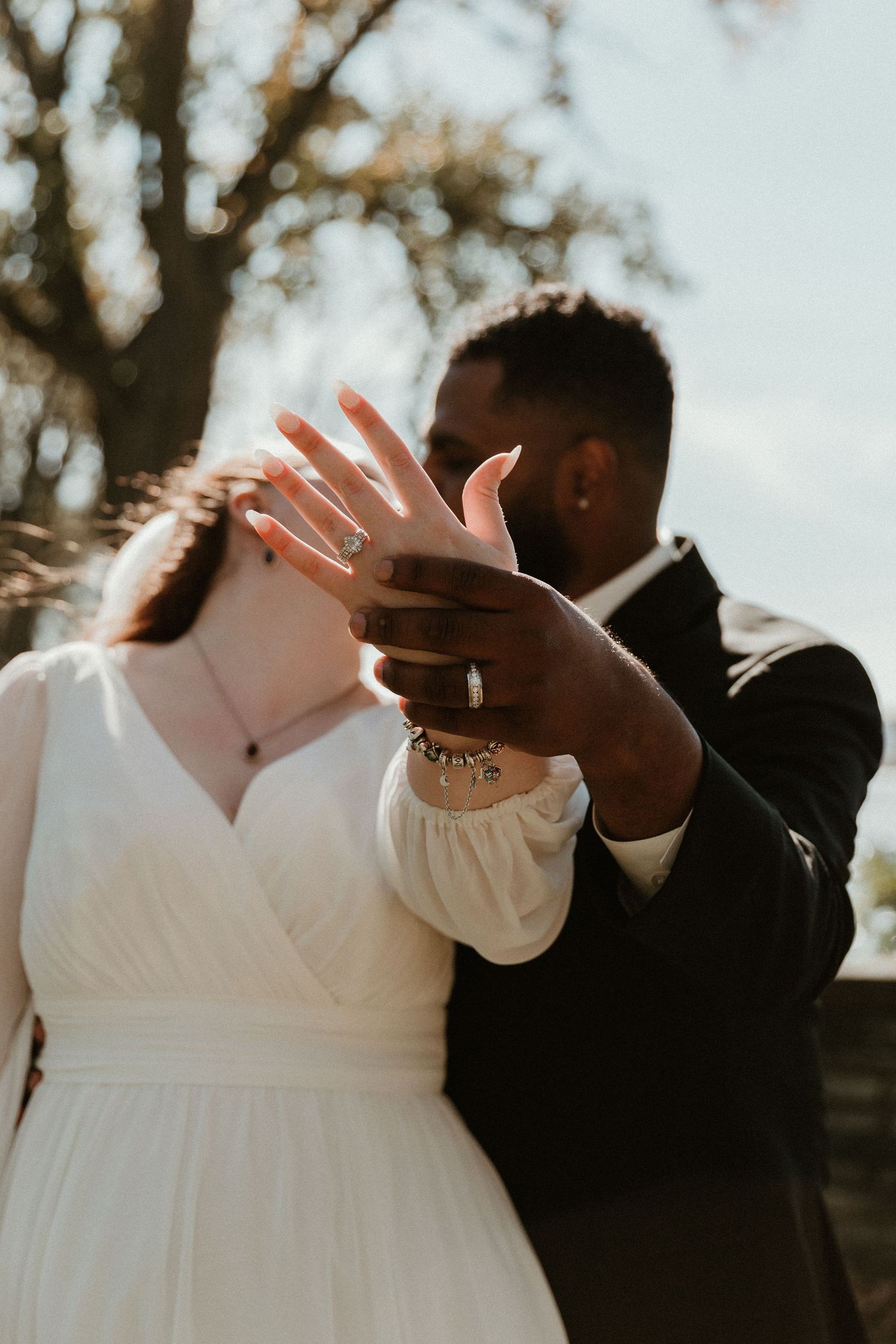 Wedding couple in white dress and black suit embrace in sunlit outdoor setting.