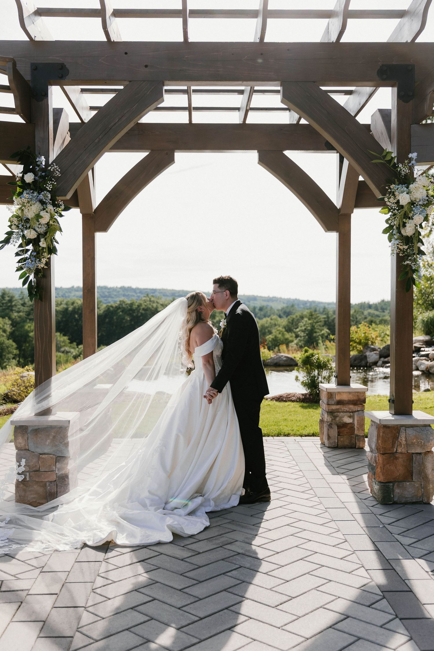 Bride and groom sharing a kiss under a wooden pergola with floral arrangements, long veil flowing, scenic outdoor venue backdrop.