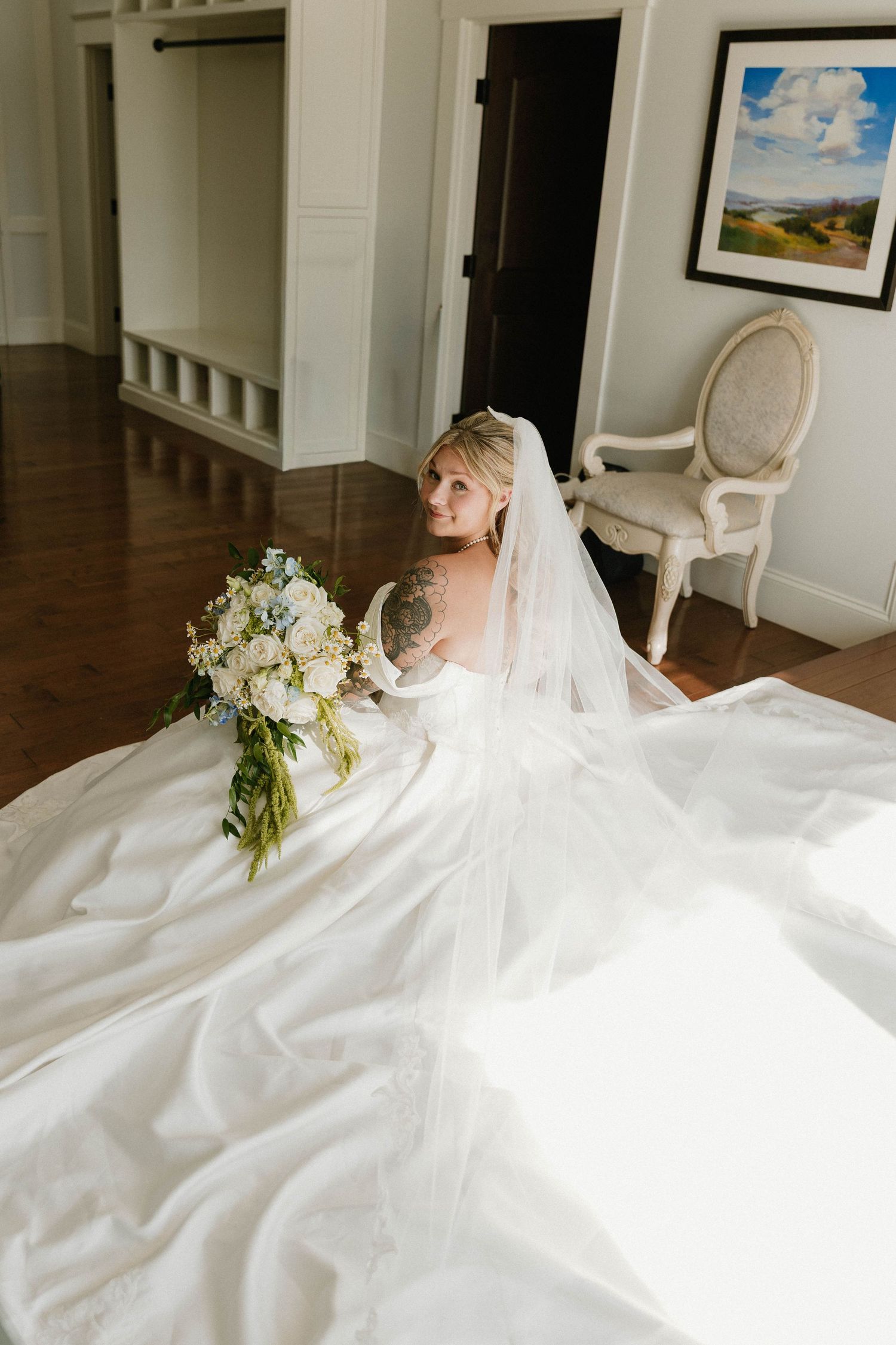 Bride in a flowing white gown and veil sits on hardwood floor holding a white bouquet in an elegant room.