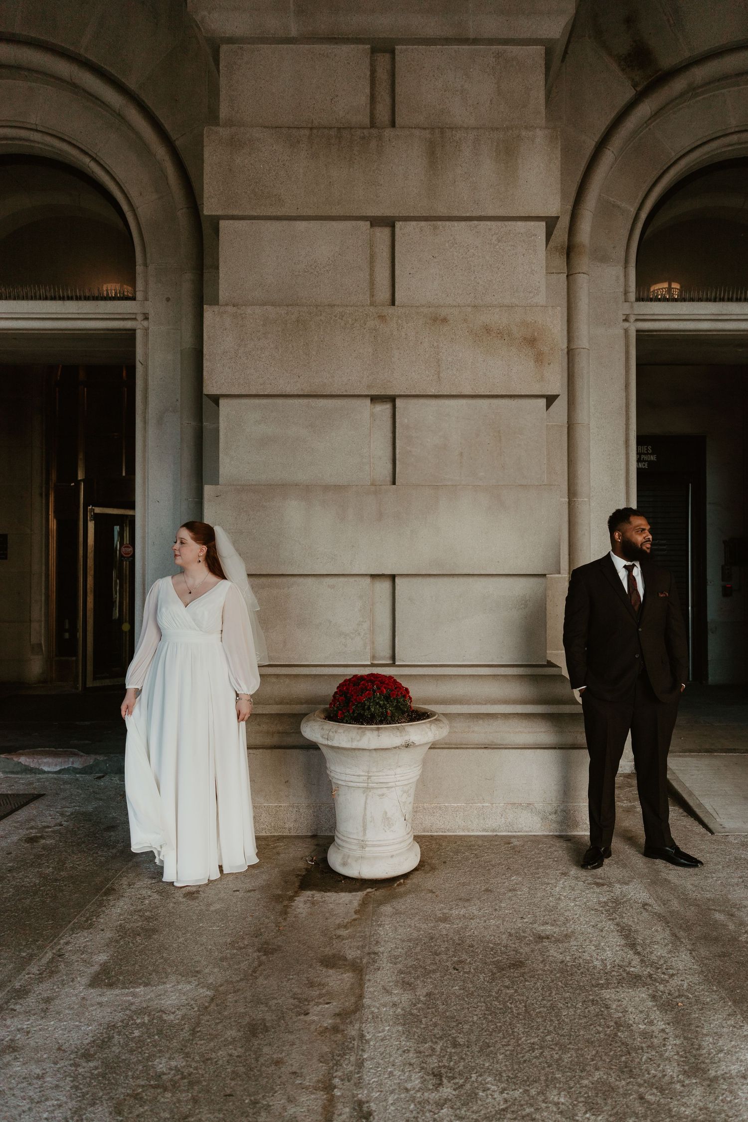 Wedding couple poses separately against stone pillars in formal attire.