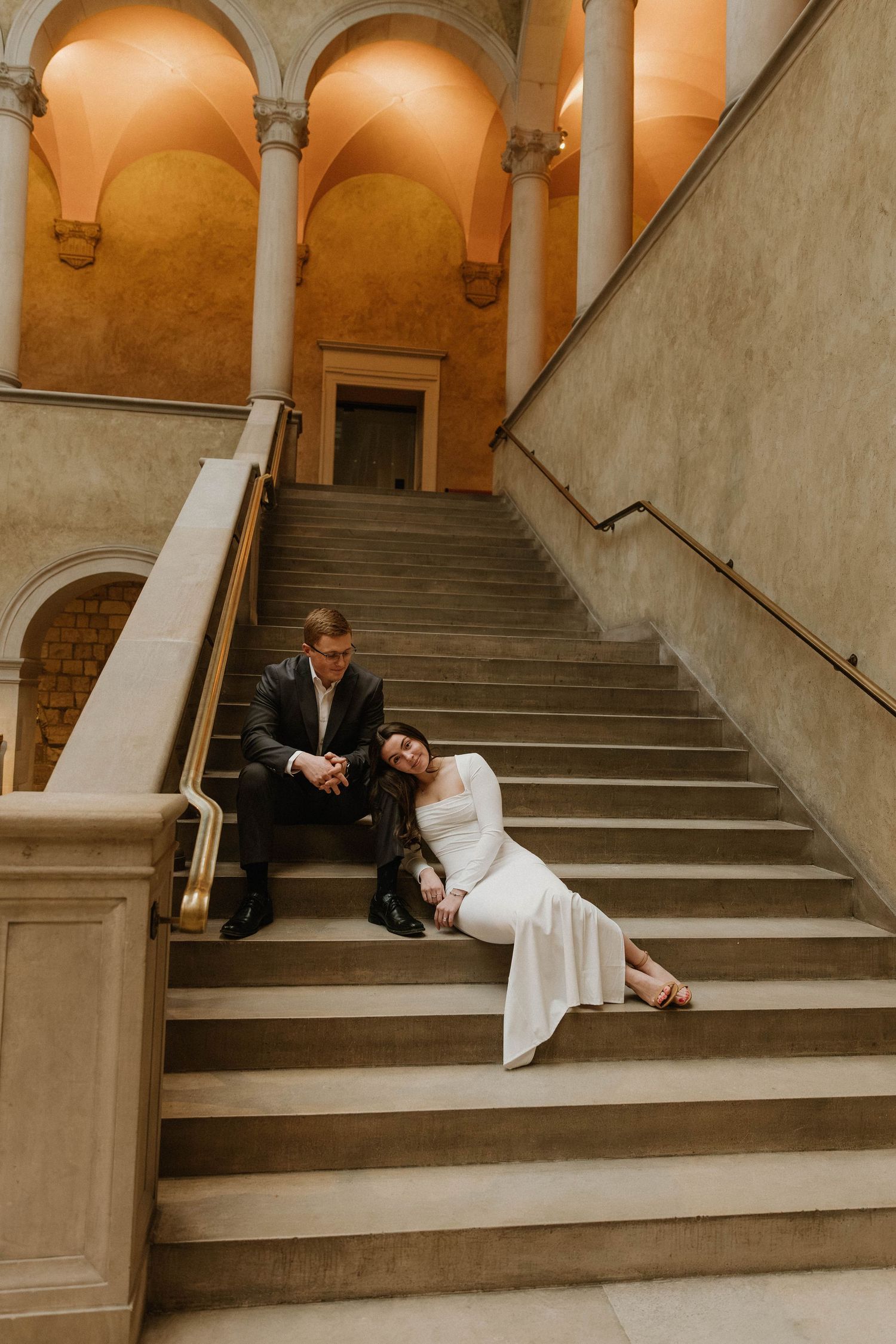 Couple sits intimately on elegant stone staircase with arched columns and warm lighting in historic building interior.
