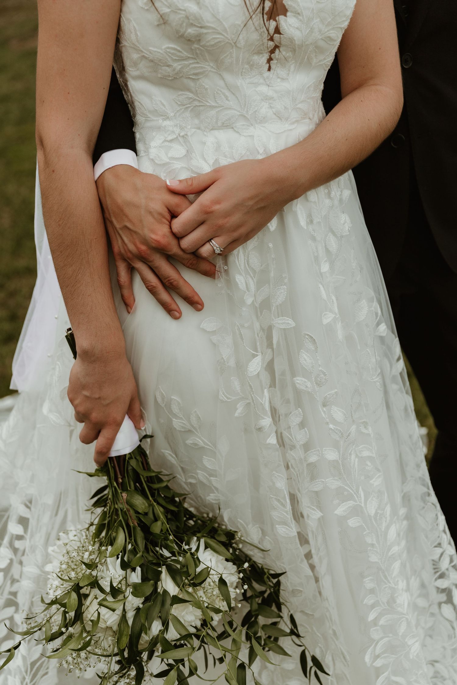 Close-up details of a white lace wedding dress with delicate floral patterns and greenery bouquet.
