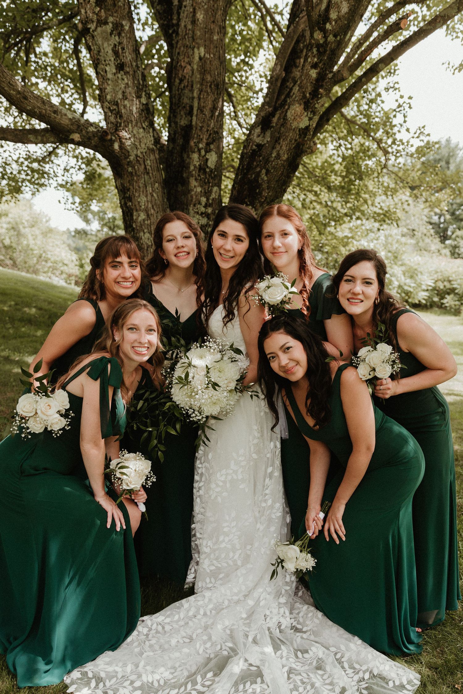 A bridal party in emerald green dresses poses together under a large tree during an outdoor wedding.