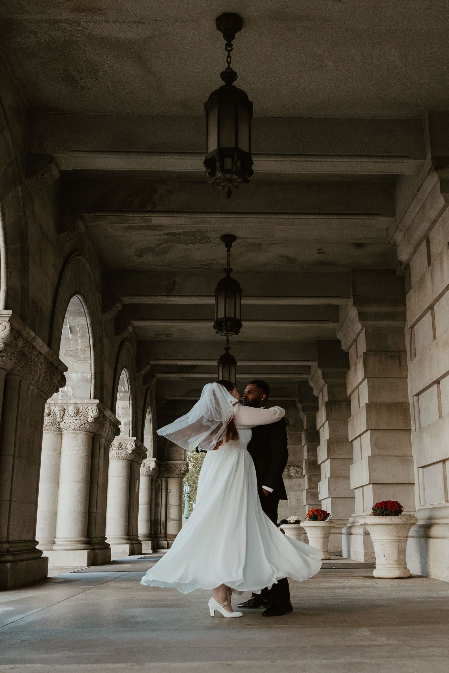 A couple dancing in an elegant colonnade with hanging lanterns and white wedding attire.