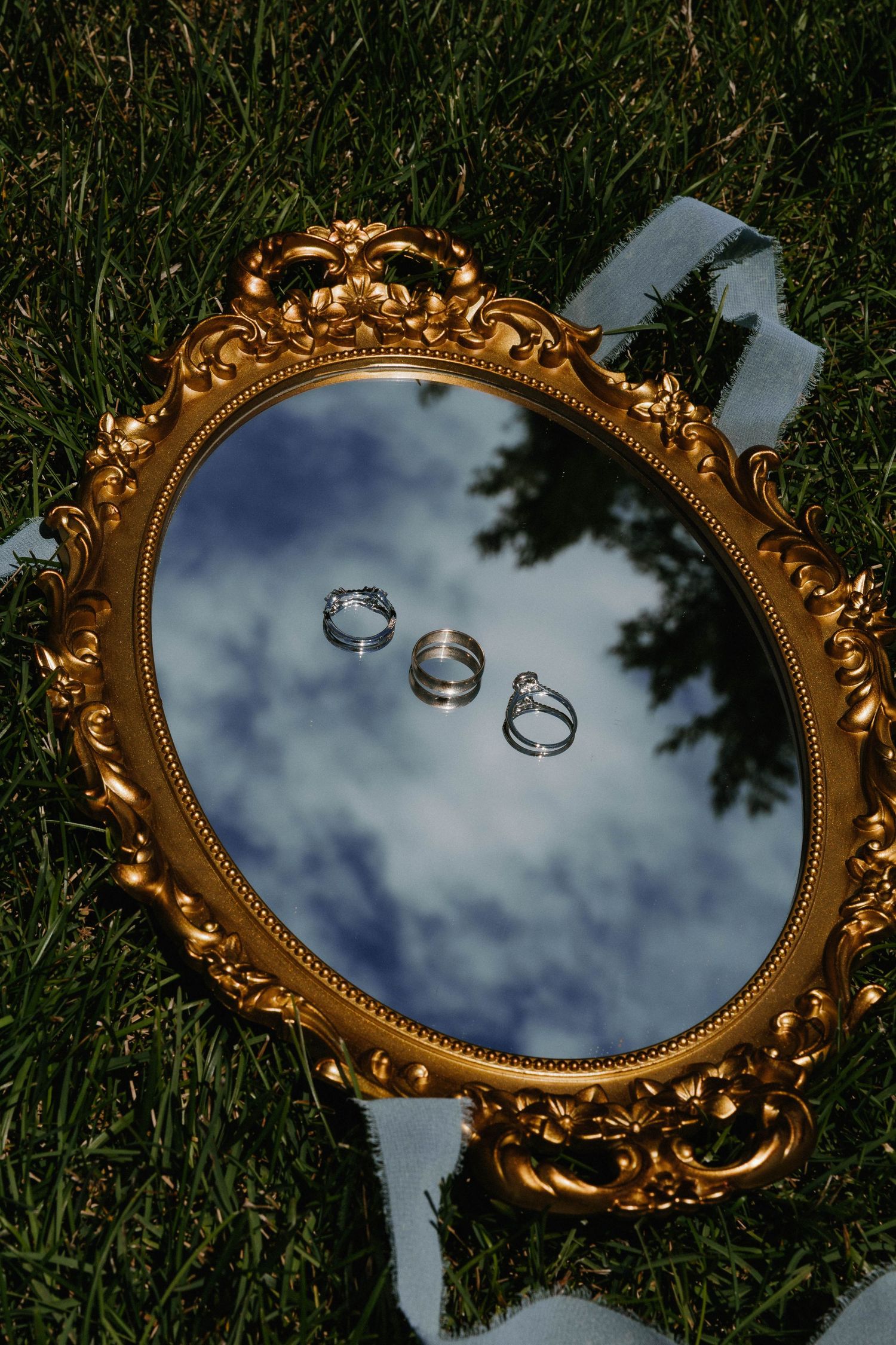 Gold ornate oval mirror on grass reflecting sky and trees, displaying three wedding rings including diamond engagement ring.