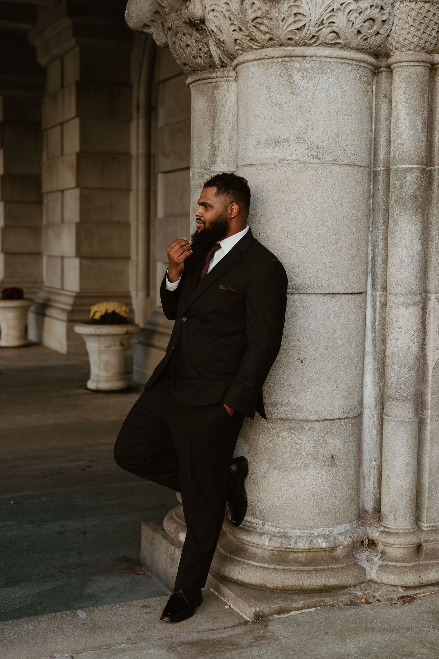 Person in formal black suit sits thoughtfully against classical stone column.