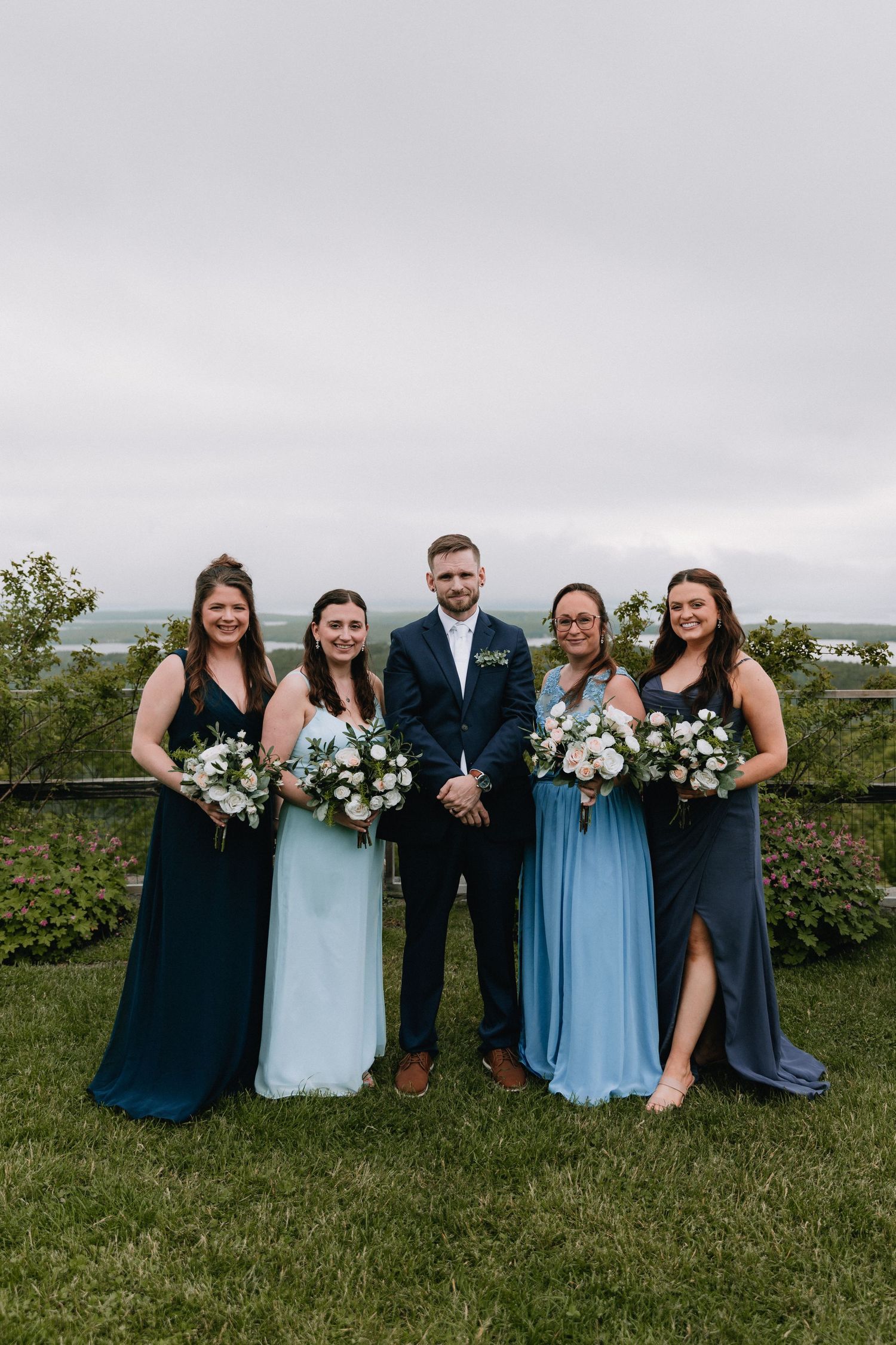 Wedding party in navy and light blue attire poses together on a scenic grassy overlook.