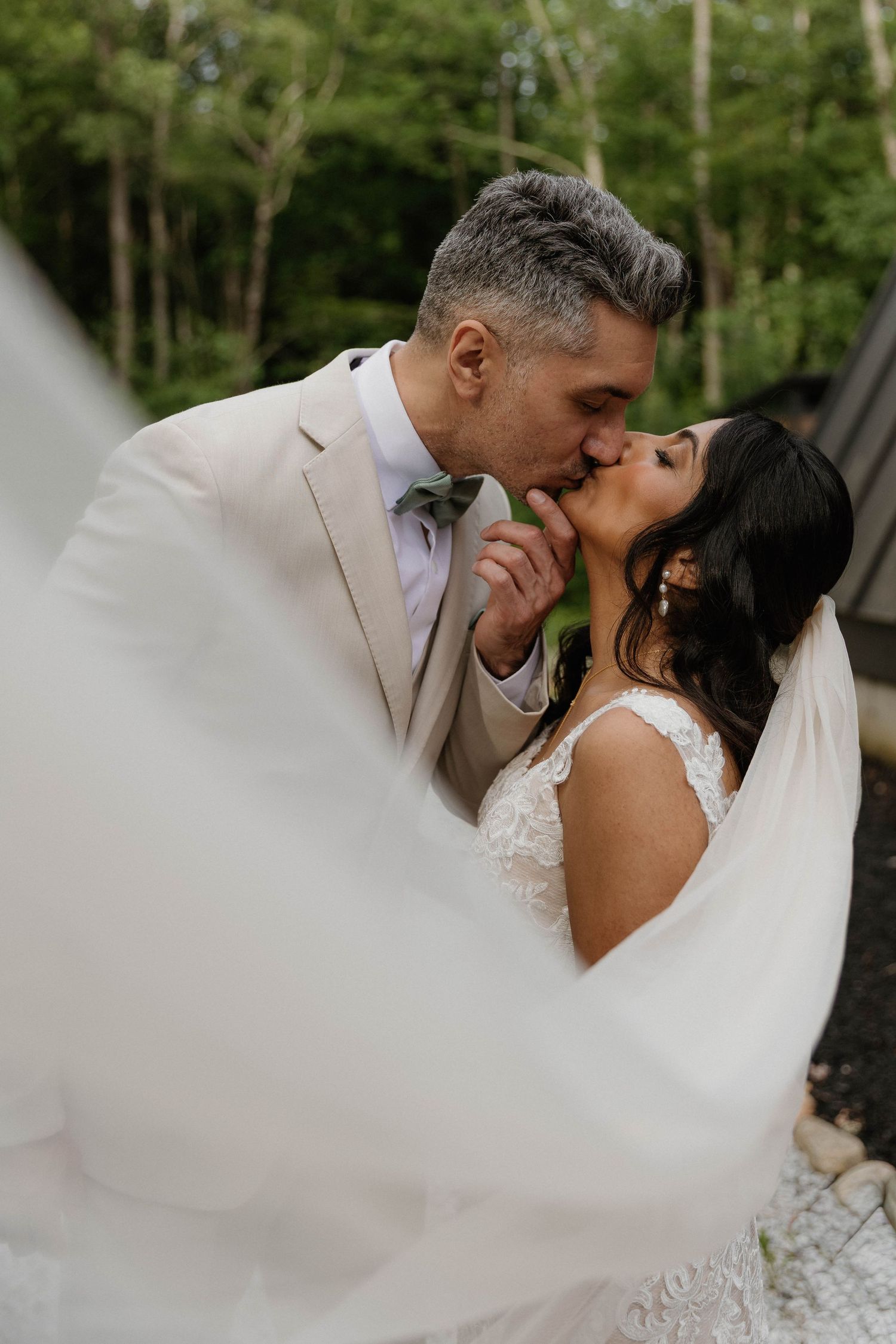 A bride and groom share a romantic kiss outdoors, with her white veil flowing in the foreground near a lush green forest.