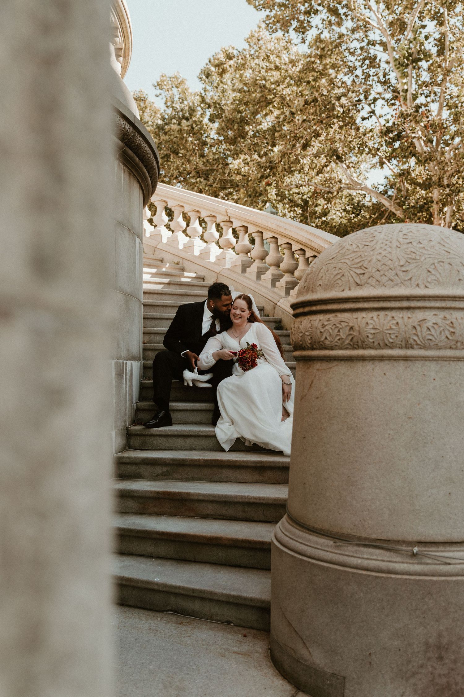 A couple in wedding attire sits on ornate stone steps during a romantic outdoor photo session.
