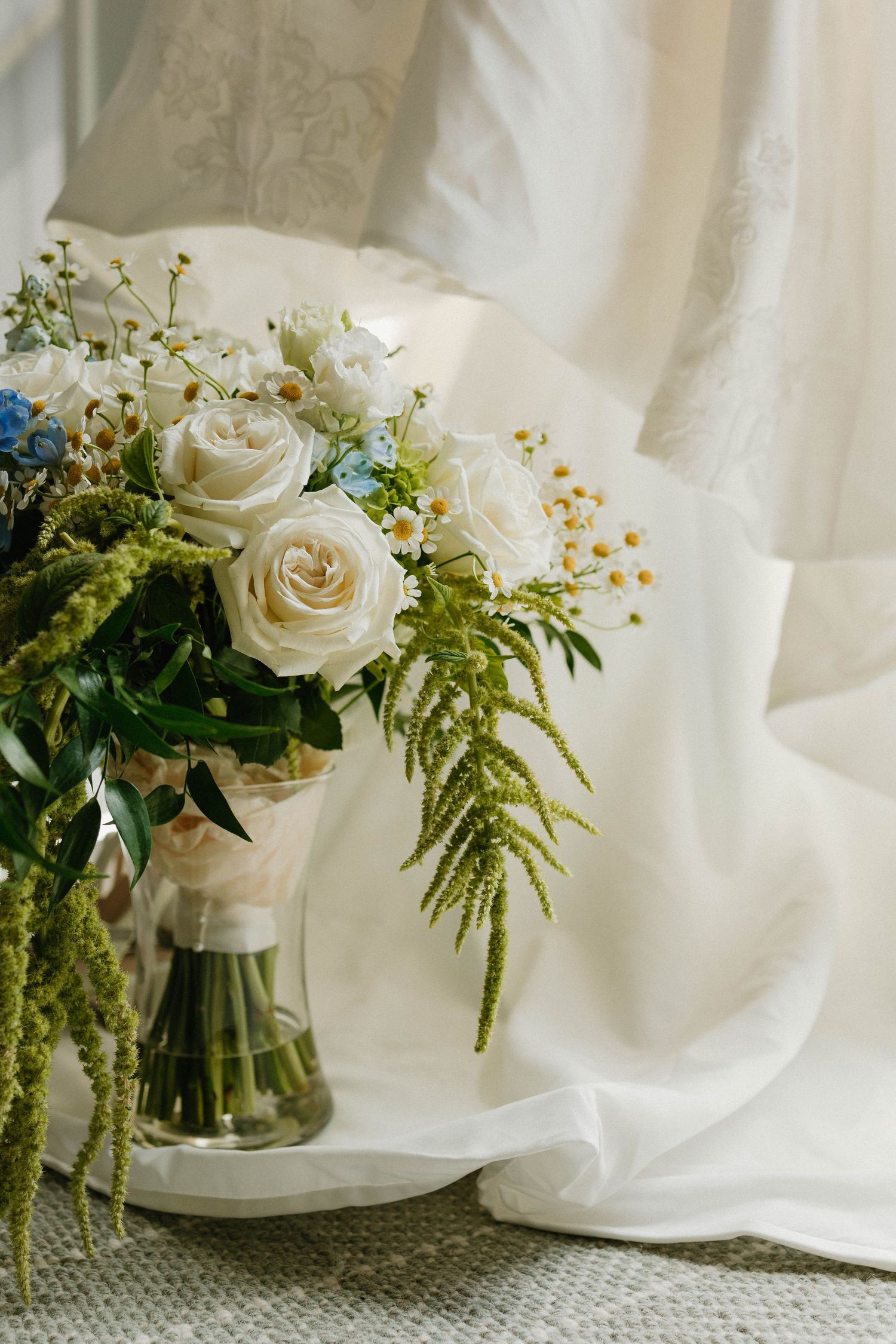 Elegant wedding bouquet with white roses, chamomile, blue flowers, and green ferns in a glass vase beside a bridal gown.