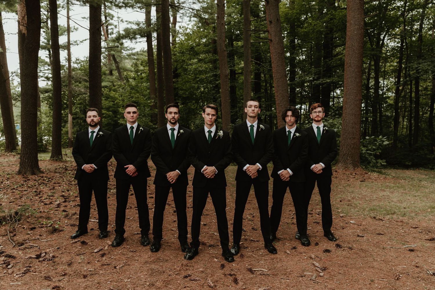 A wedding party in black suits stands together in a wooded forest setting for a group photo.