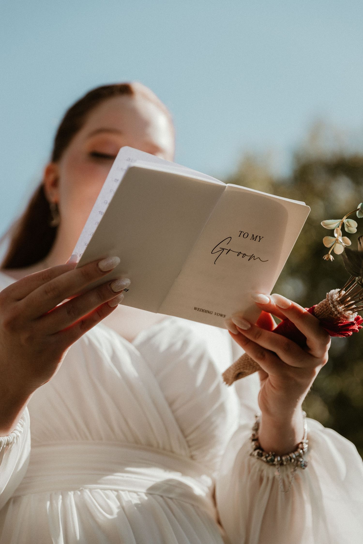 A bride in white holds a book while sitting outdoors on a sunny day.