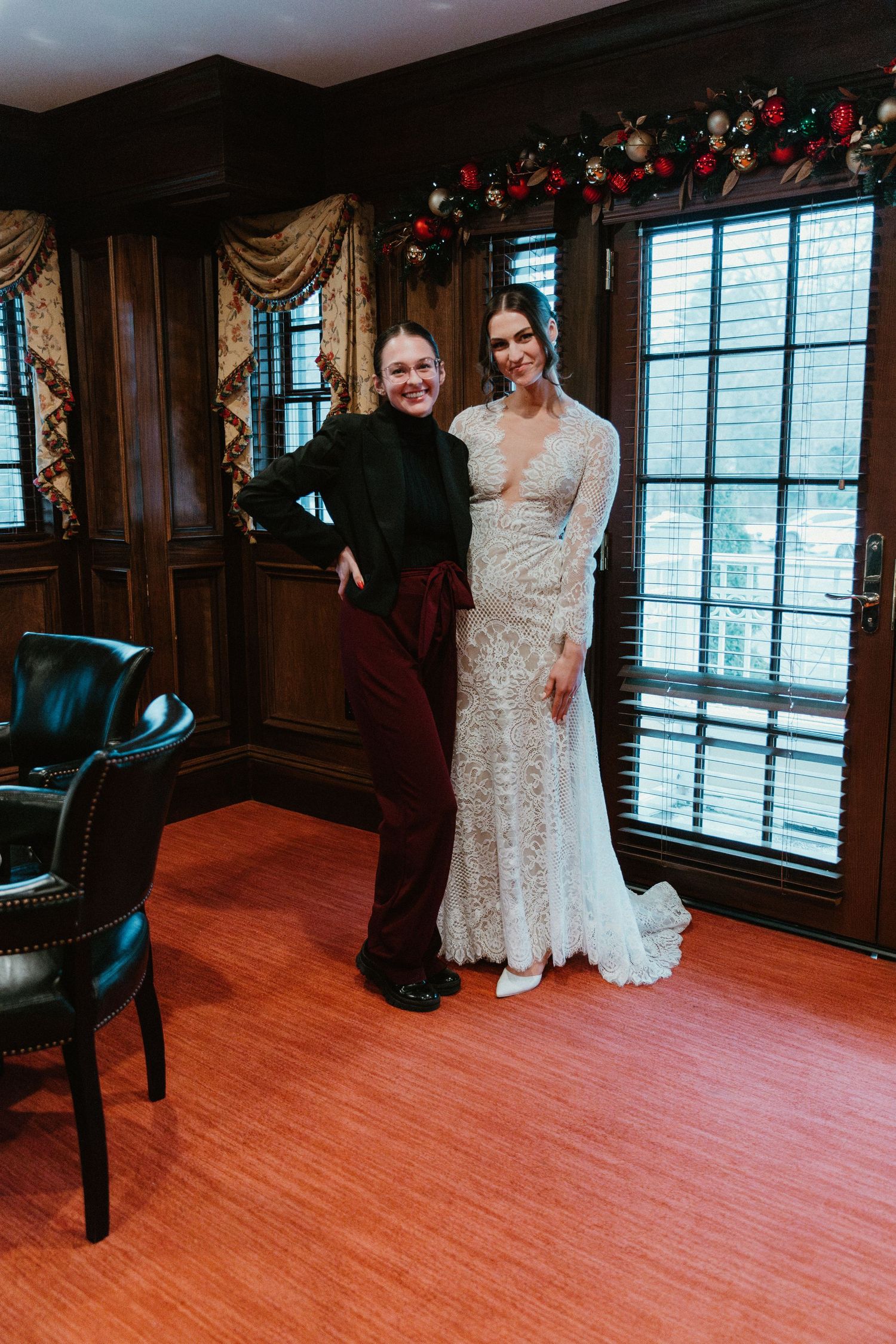 Two people in elegant white lace wedding dresses share a joyful moment on a hardwood floor in an ornate room.