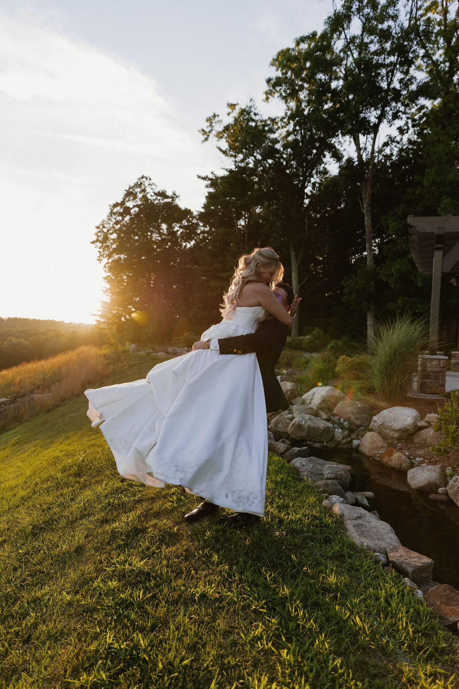 Groom lifting bride in white gown at sunset near a pond, surrounded by lush greenery on their wedding day.