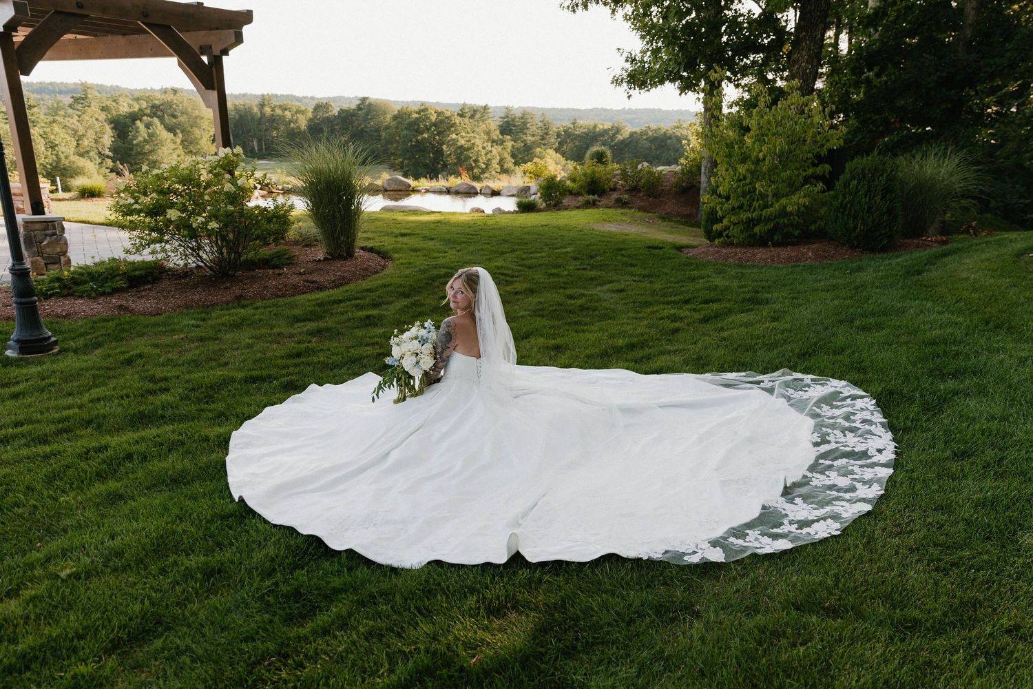 Bride in a stunning white ball gown with long train sits on lush green lawn holding a bouquet at an outdoor wedding venue.