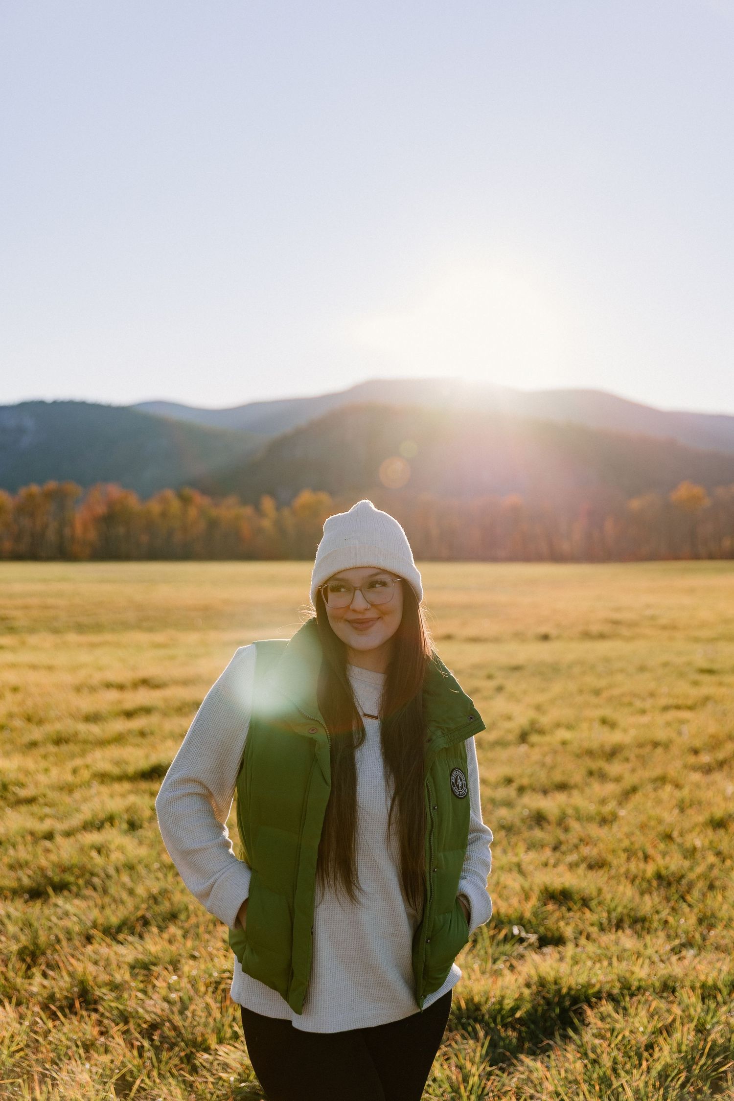 Person in white beanie and green vest poses in golden field at sunset with mountains in background.