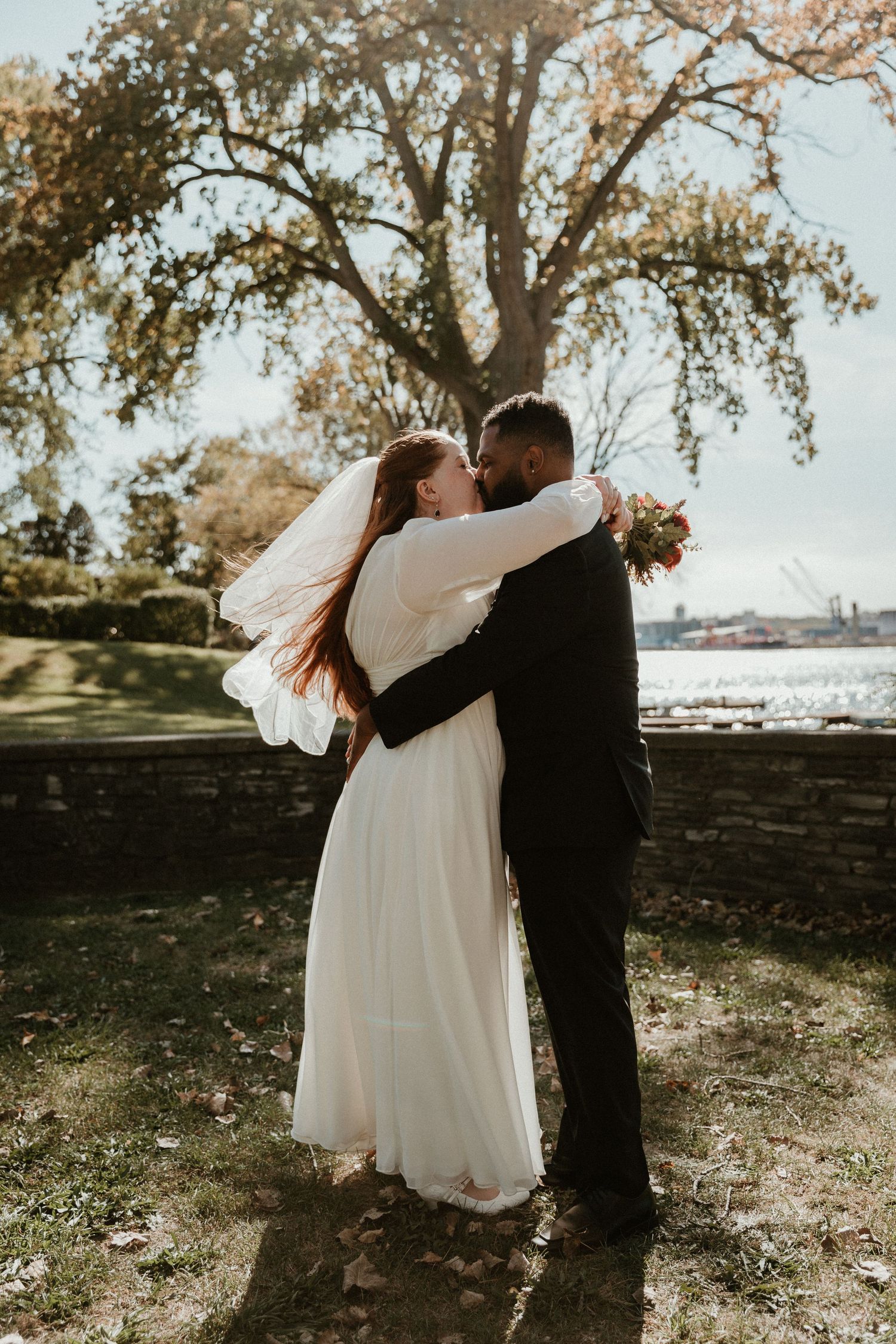 A wedding couple embraces under a large tree with autumn foliage and waterfront view.