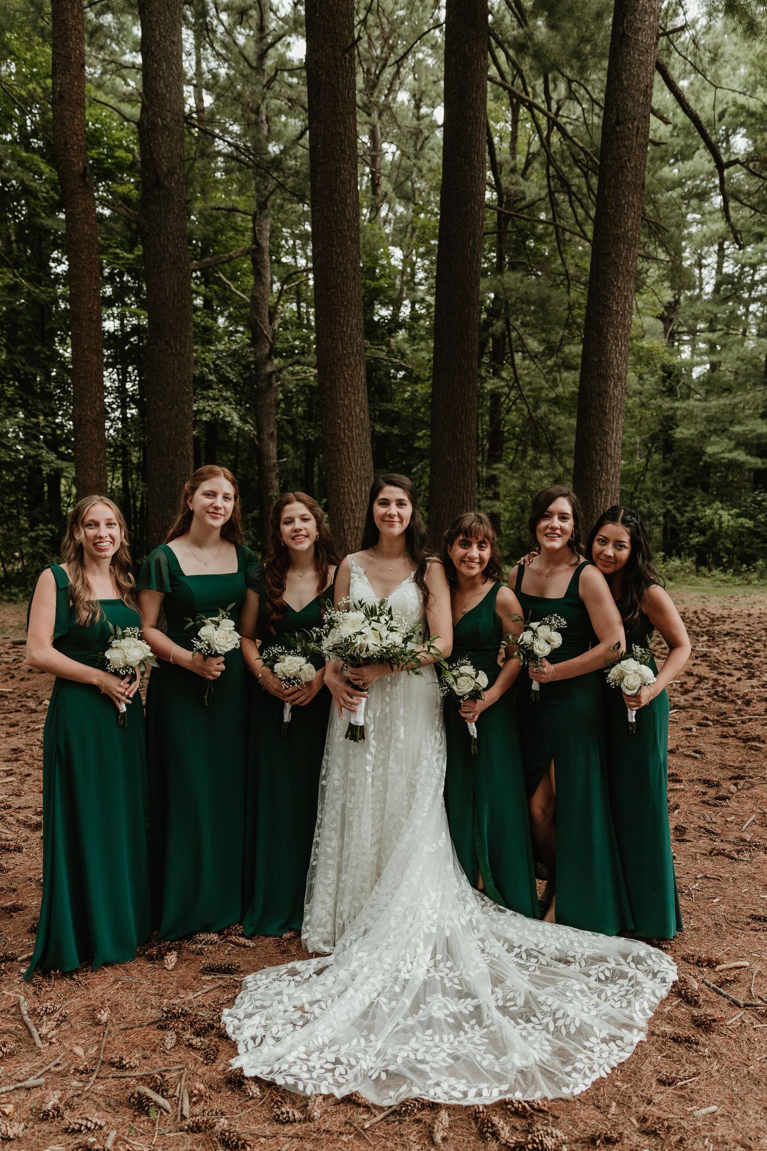 Wedding party with bridesmaids in emerald green dresses and bride in lace gown pose in forest setting.