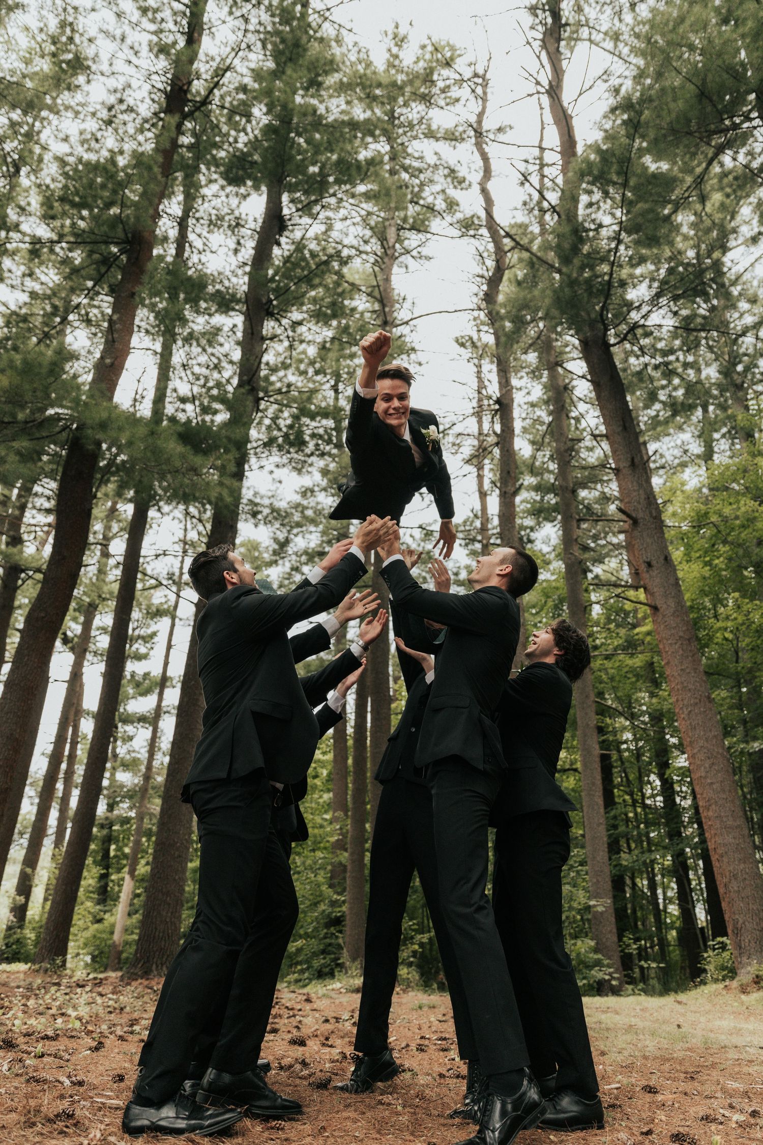Groomsmen in black suits posing playfully in wooded outdoor setting.