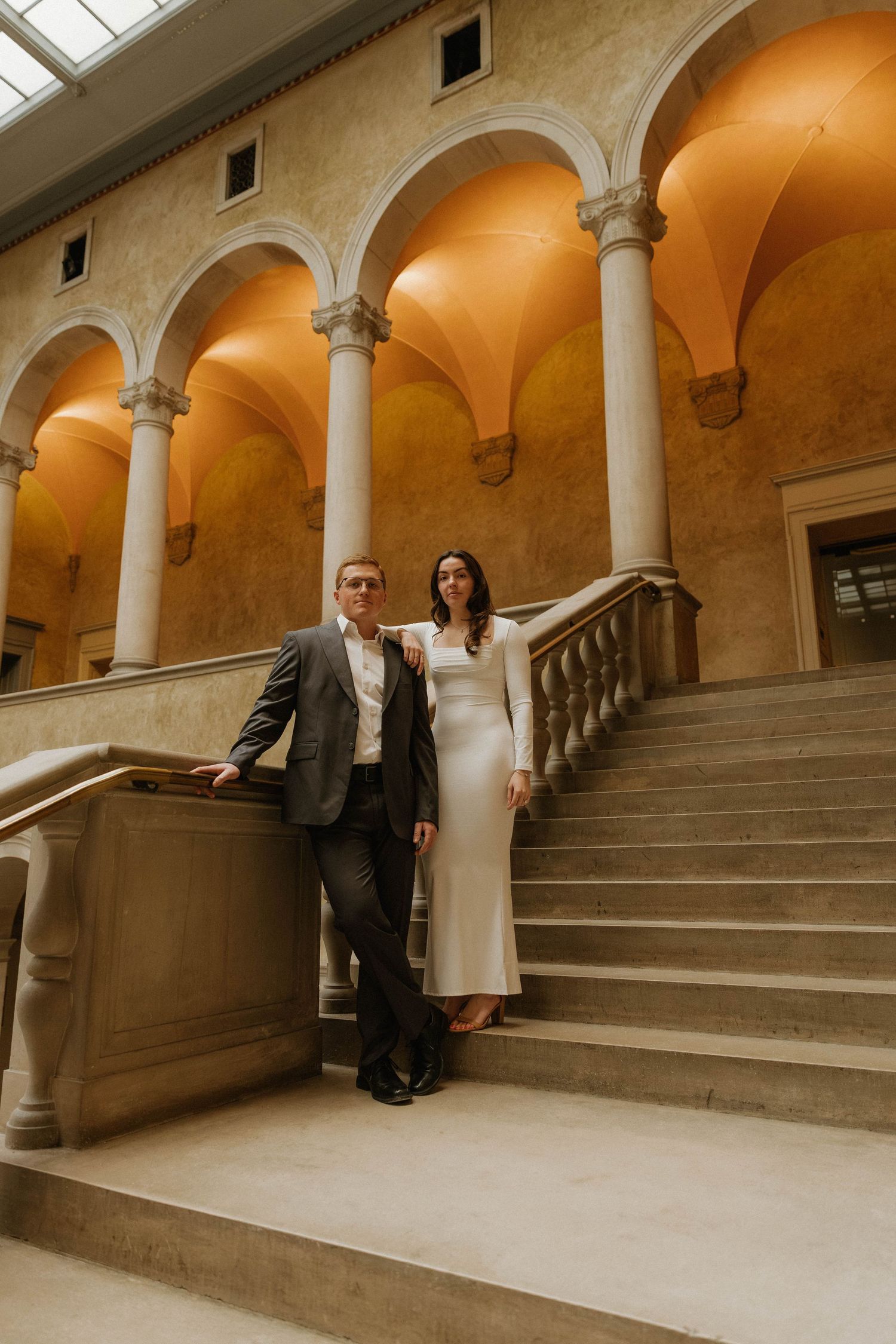 Elegant couple in formal attire pose on grand marble staircase in historic building with arched colonnade.