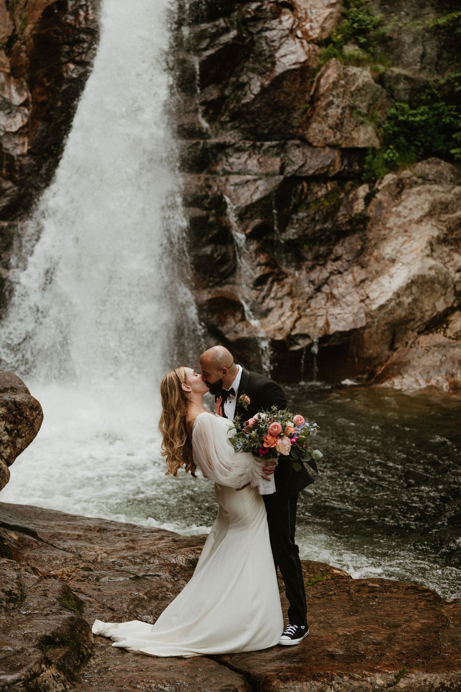 A romantic wedding photo session in front of a majestic waterfall with a flowing white dress and dark suit.