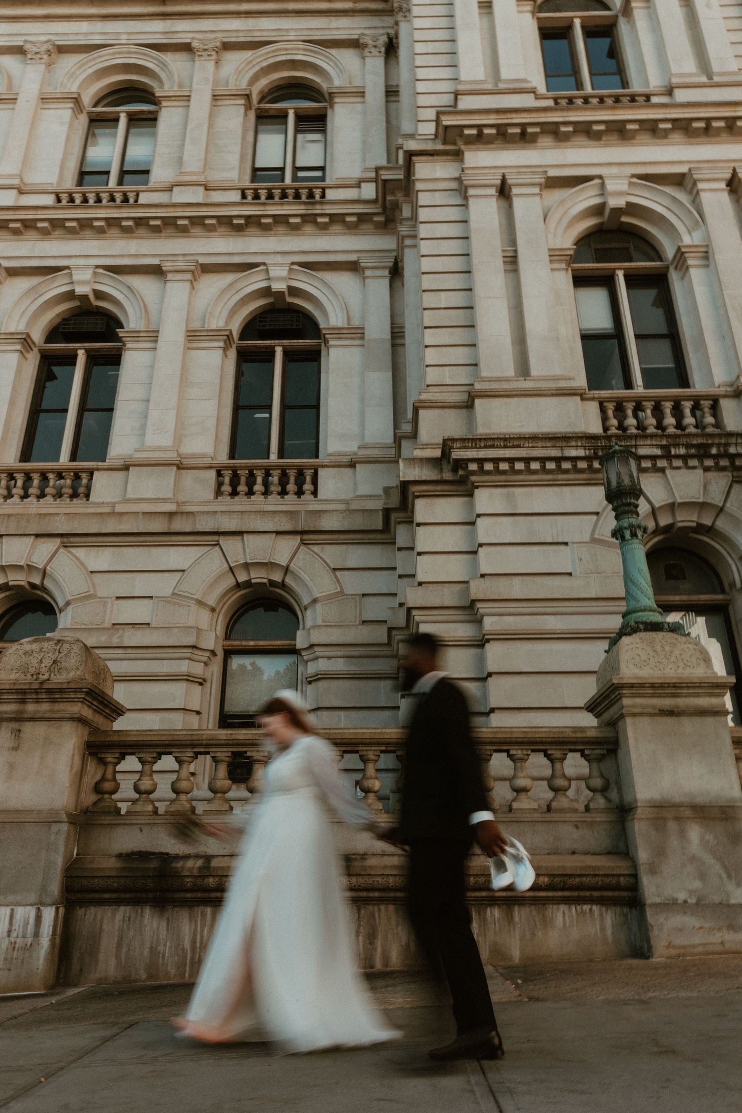 A couple in wedding attire walks past a grand historic building with arched windows.