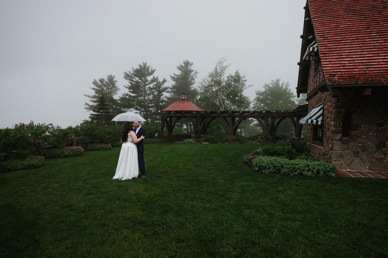 A wedding couple poses on a grassy hill near stone buildings with umbrellas in the distance.