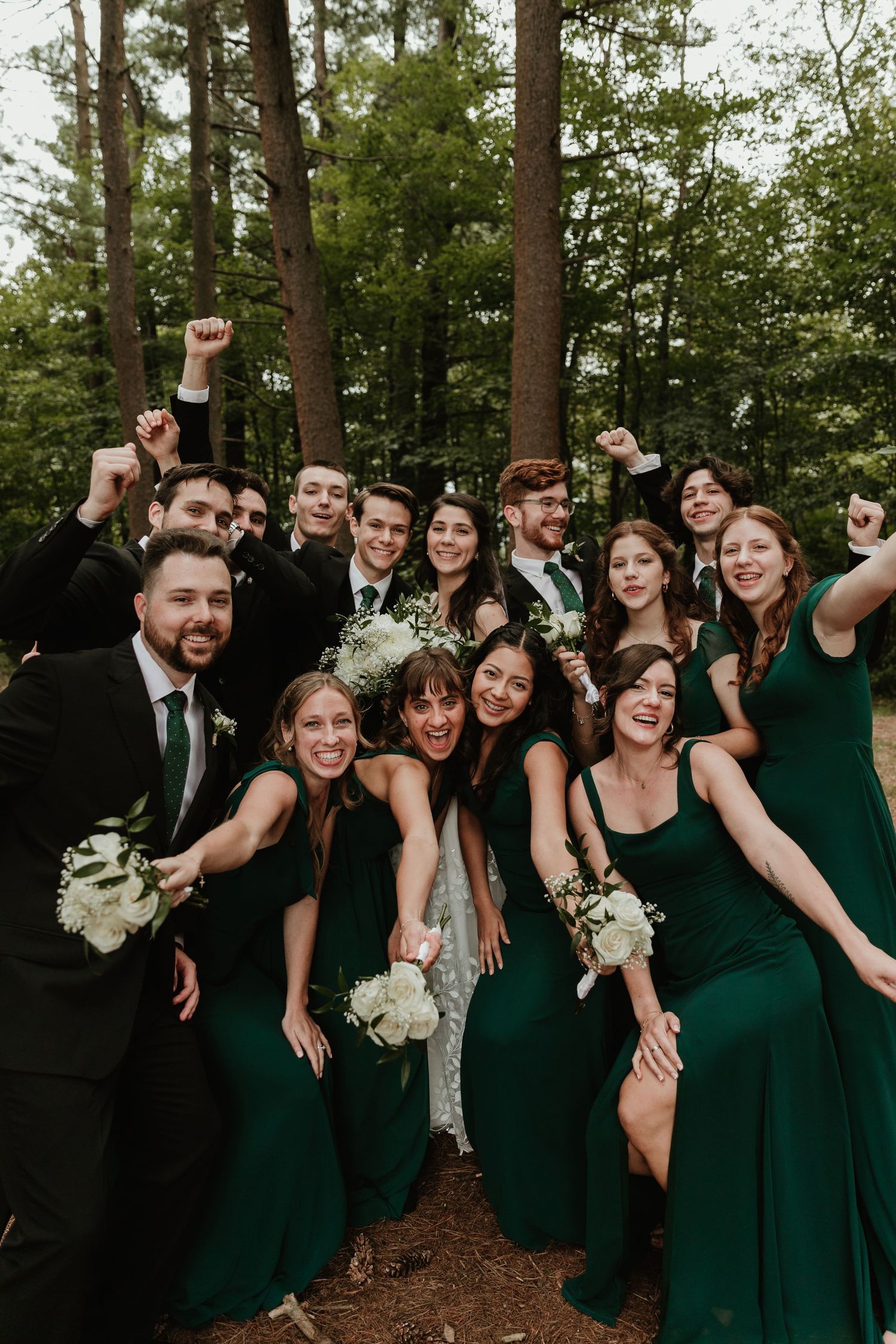 Wedding party in emerald green dresses and black suits pose playfully together in a forest setting.