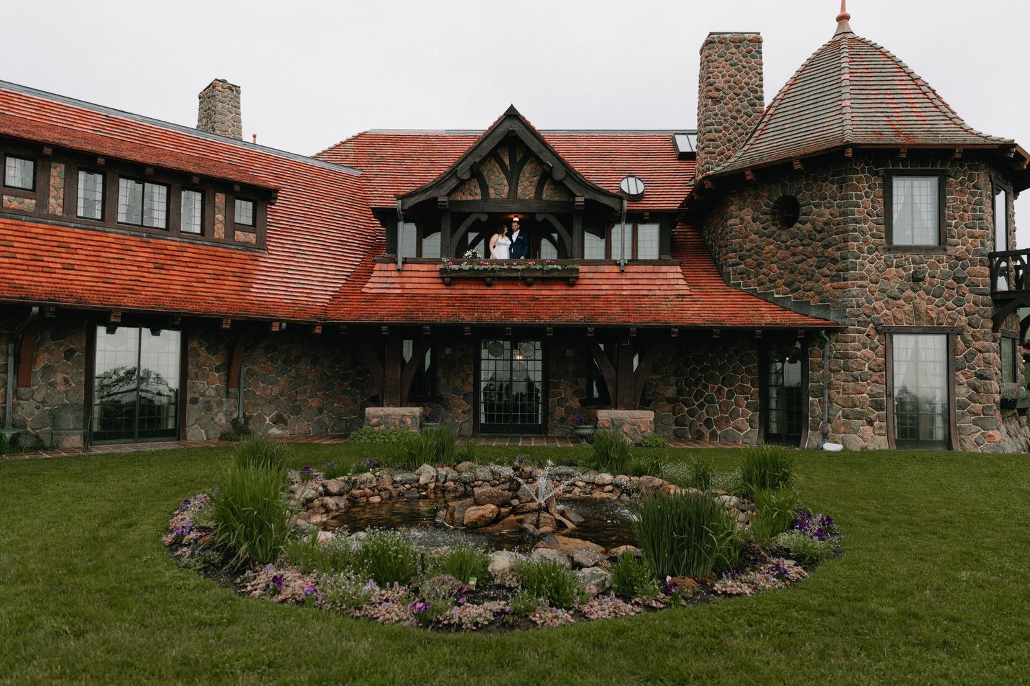 Stone mansion with red roof and Tudor-style architecture set against green landscape.