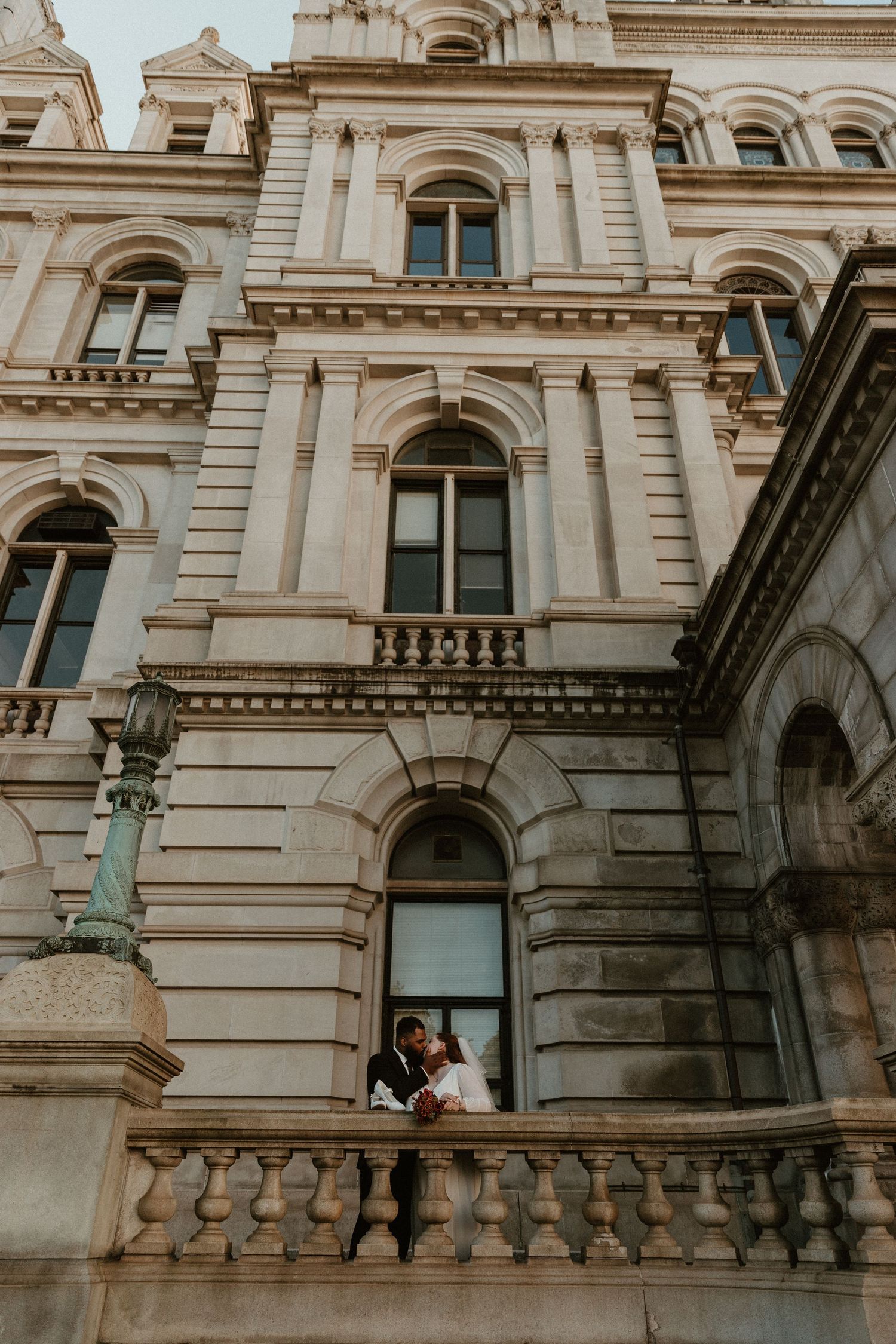 A wedding couple poses on a balcony of a grand historic building with ornate architecture.