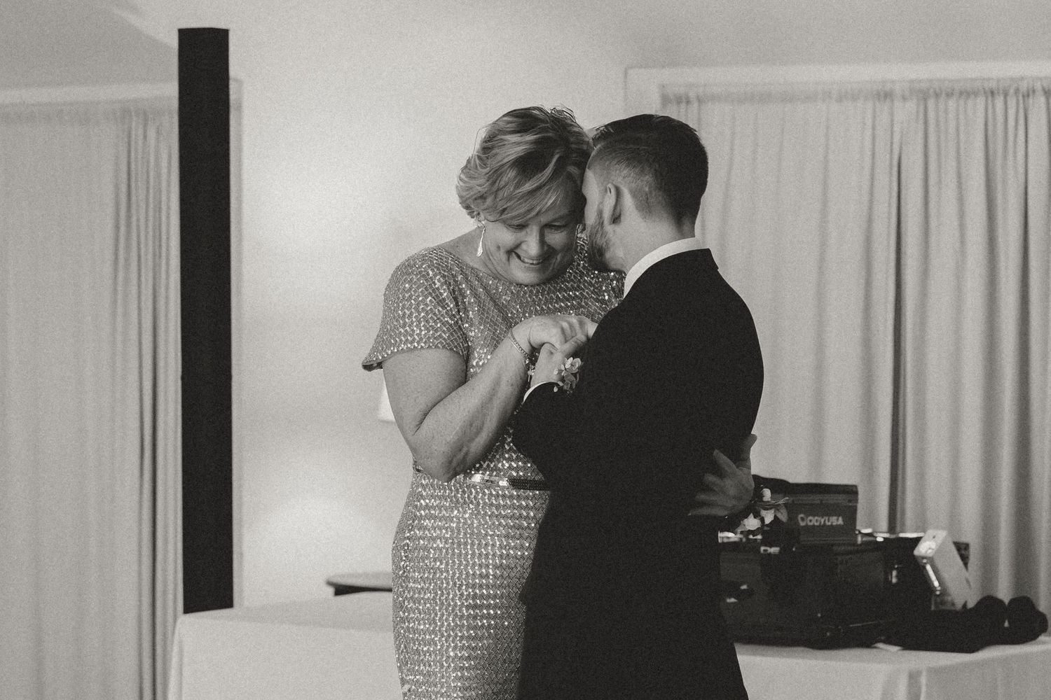 A black and white photo captures an intimate dance moment at an indoor wedding reception.