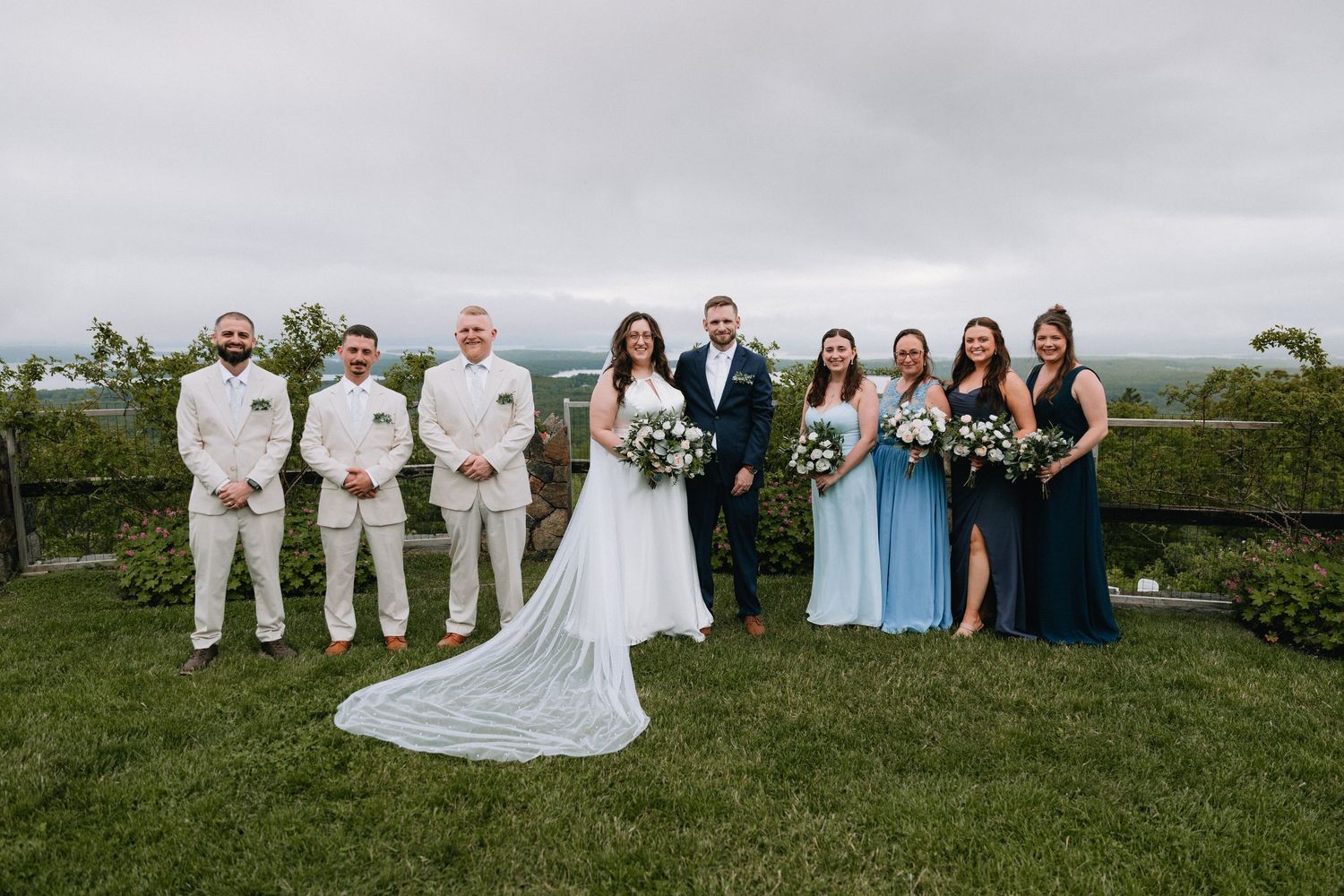Wedding party in white, navy, and light blue attire poses on grassy hillside for group photos.