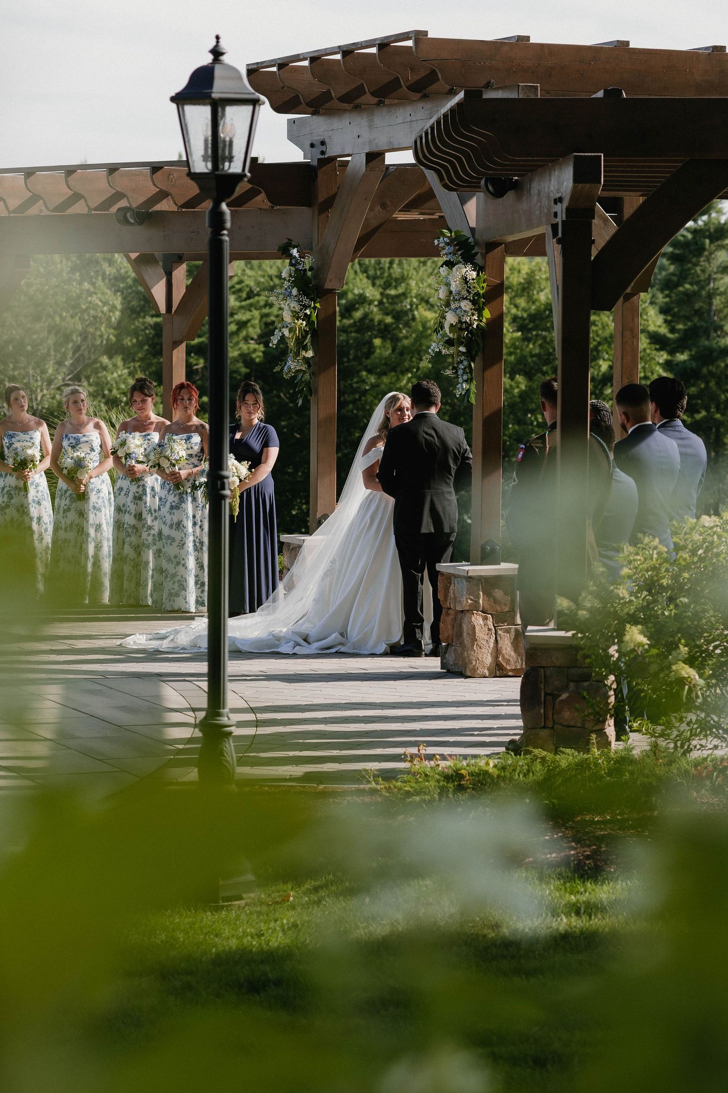 Bride and groom exchange vows under a wooden pergola outdoors, with bridesmaids in navy dresses standing nearby.