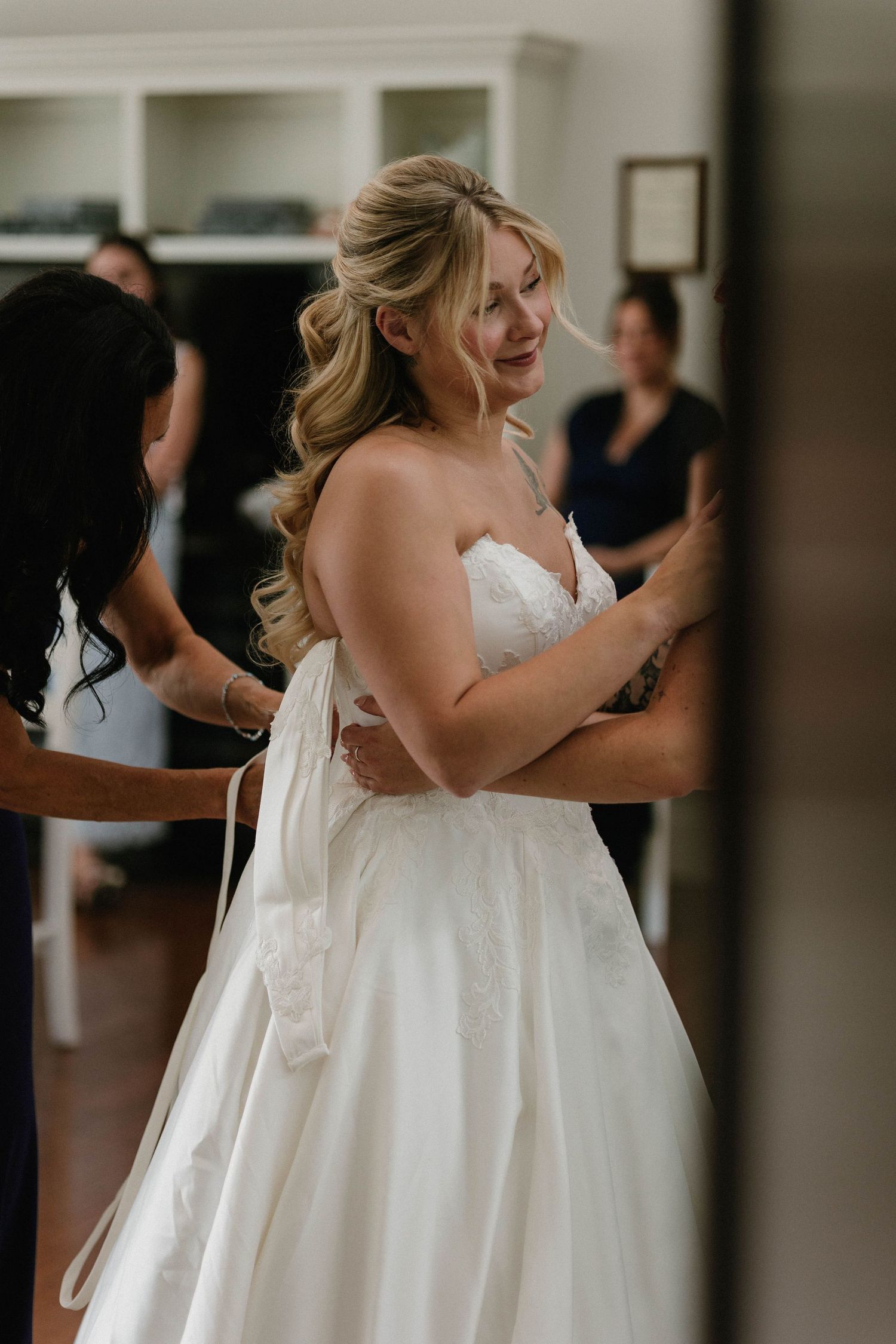 Bride in white strapless wedding gown being helped by bridesmaids while smiling over her shoulder.