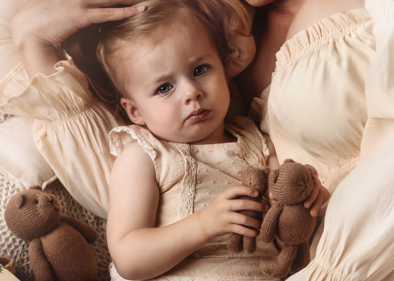A small child rests on cream-colored bedding while holding brown teddy bear toys in a vintage-style portrait.