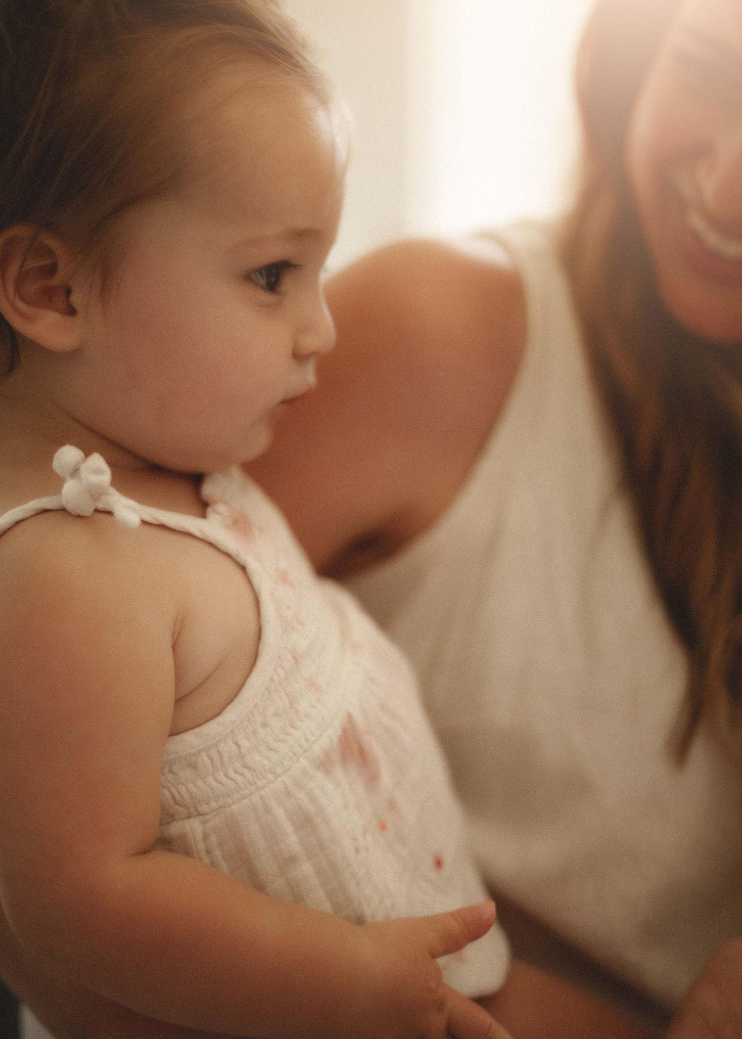 Profile view of a small child wearing a white sleeveless dress in soft warm lighting.