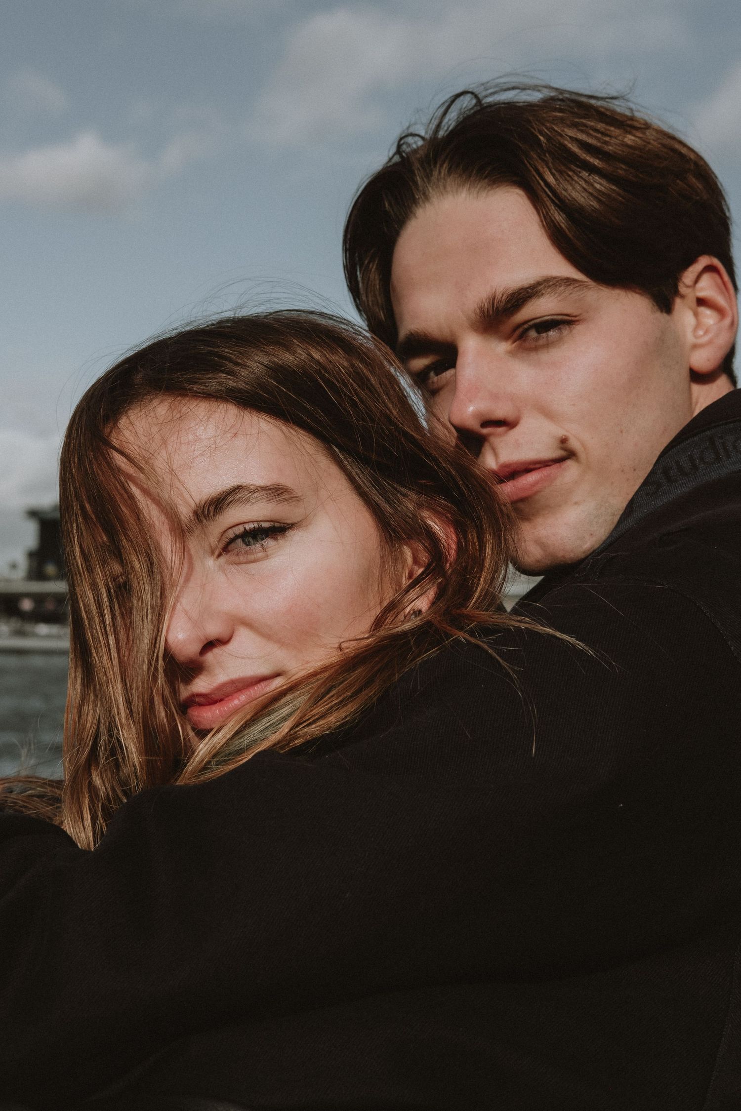 Young couple embracing outdoors near waterfront, woman leaning head on mans shoulder under cloudy sky.