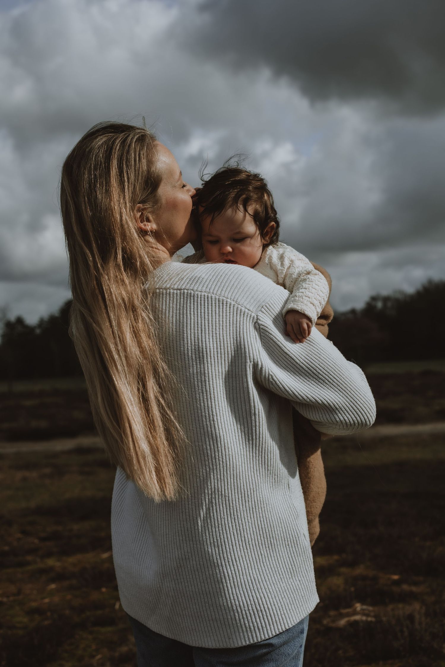 A loving mother kisses her baby tenderly under a dramatic cloudy sky.