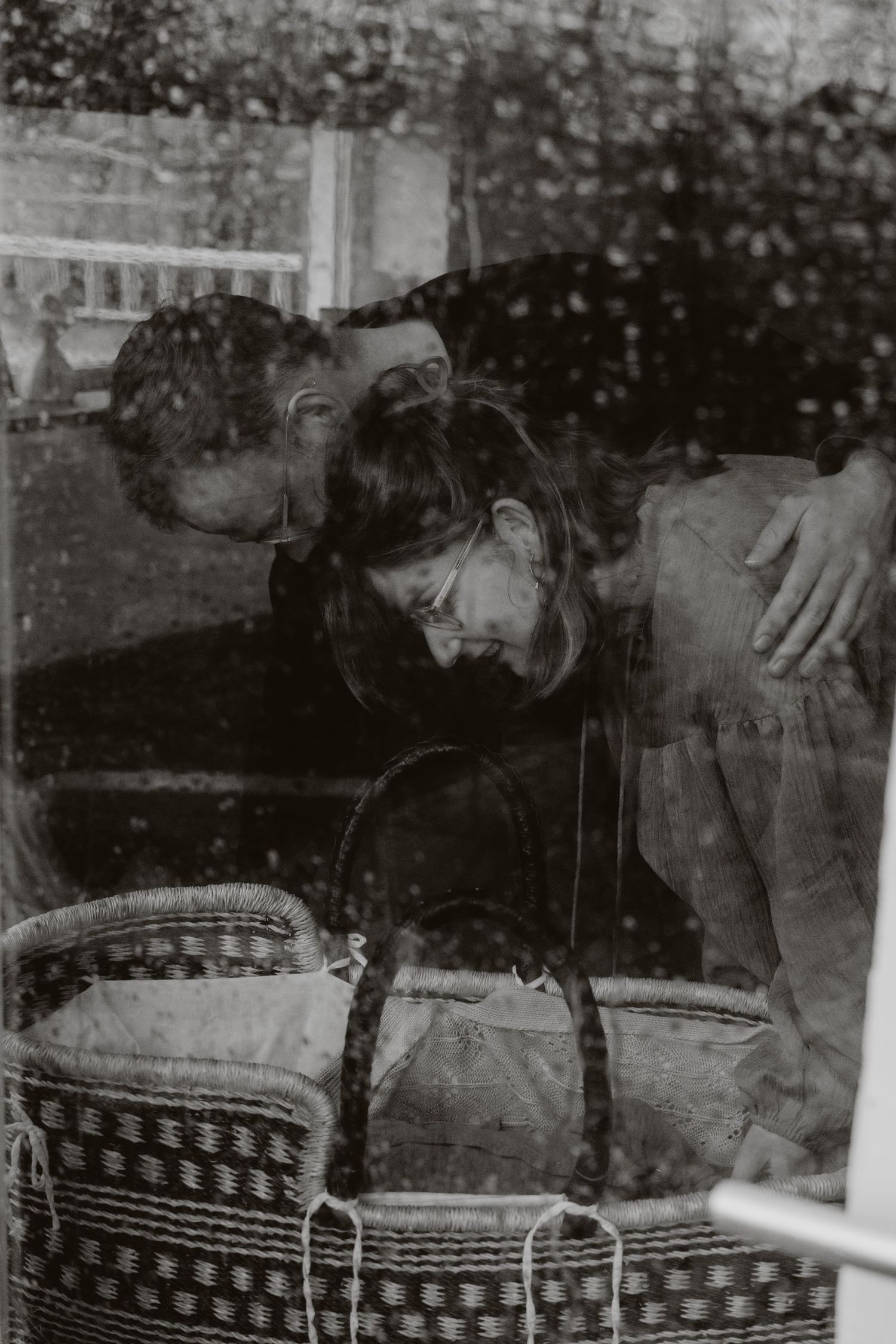 Black and white photo of couple gazing at newborn in wicker bassinet through rainy window.
