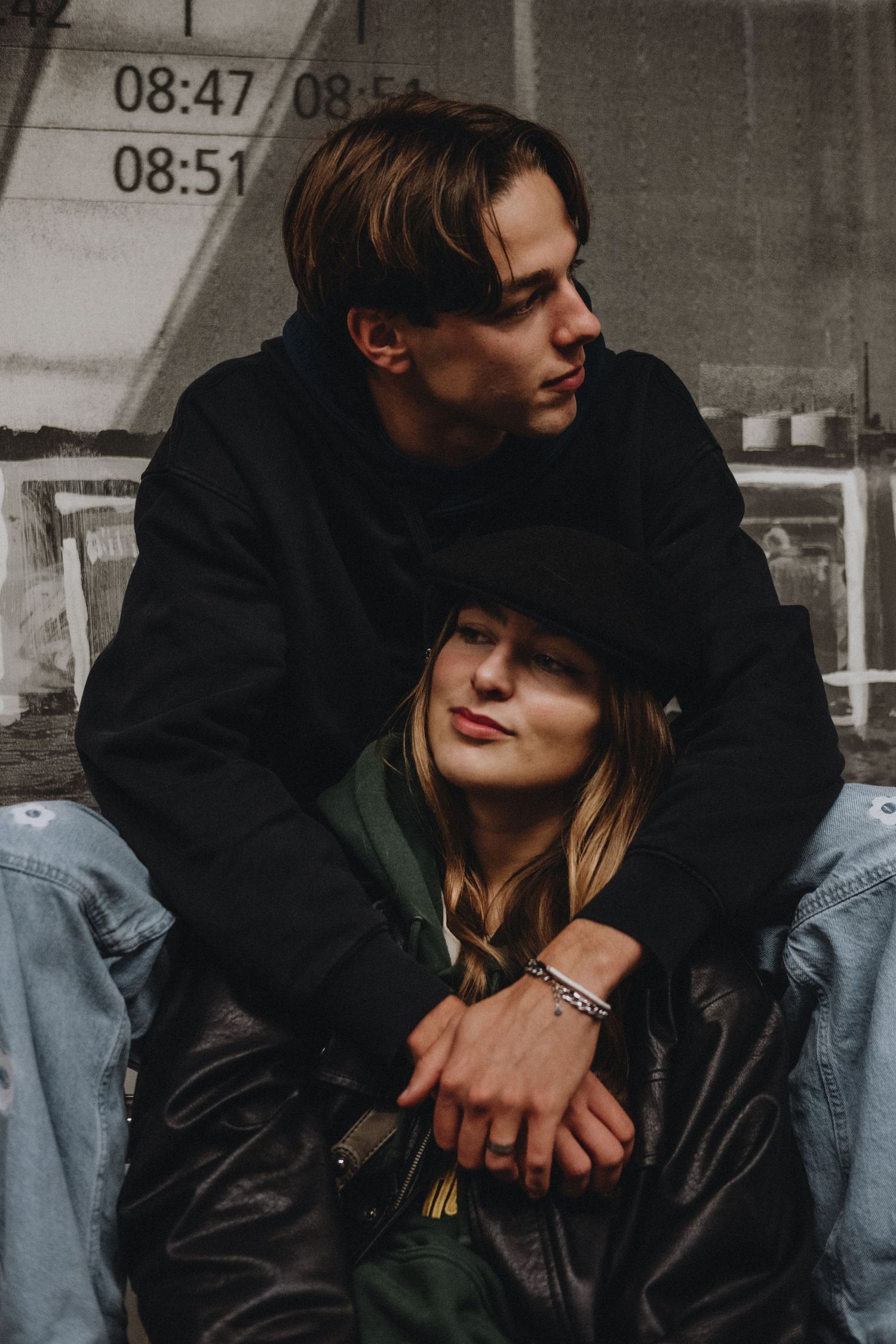 Intimate portrait of a young couple sitting together, man embracing woman against an urban wall.