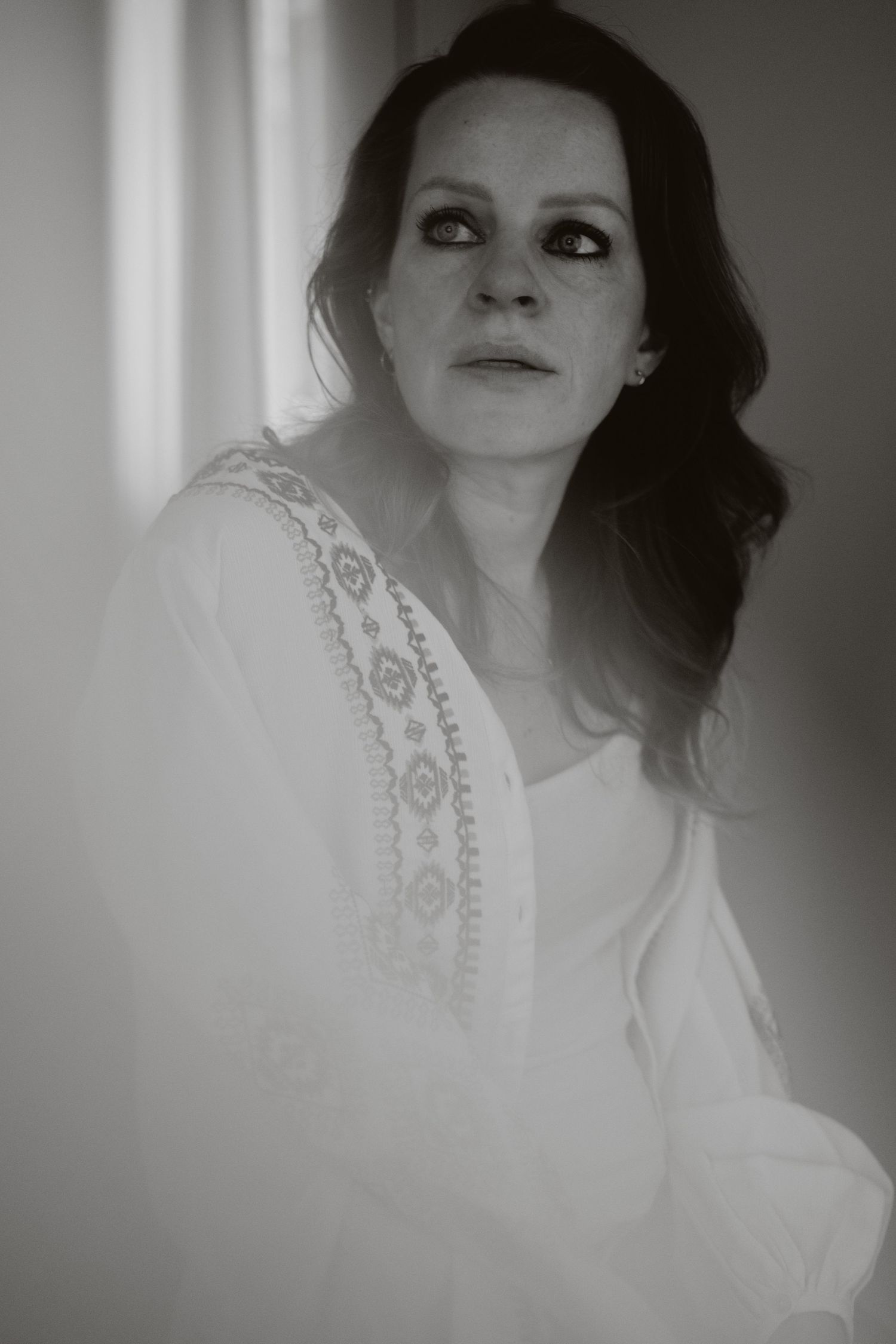 Dreamy black and white portrait of a woman in embroidered white dress near a window.