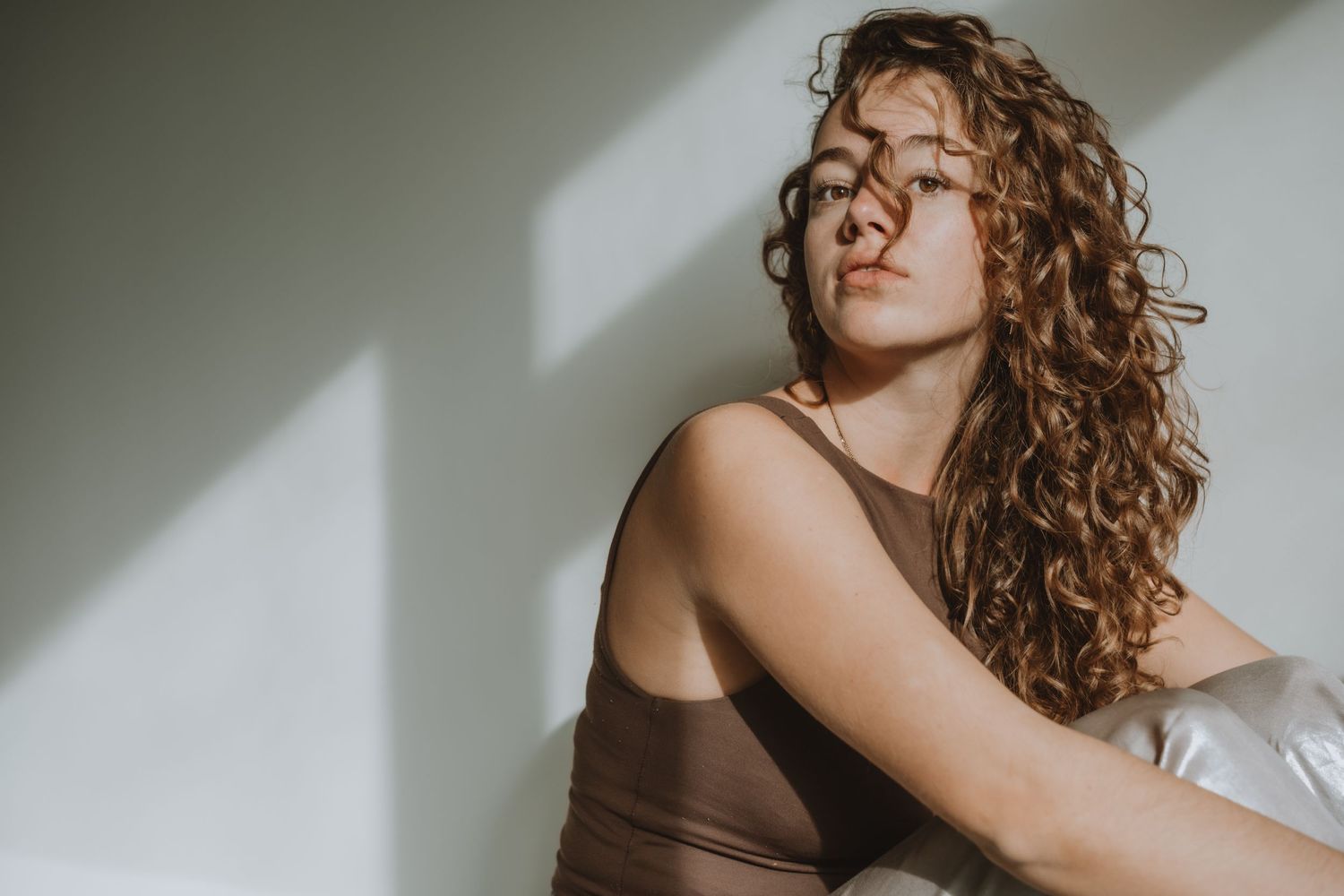 Young woman with curly auburn hair relaxing in warm sunlight against a white wall.