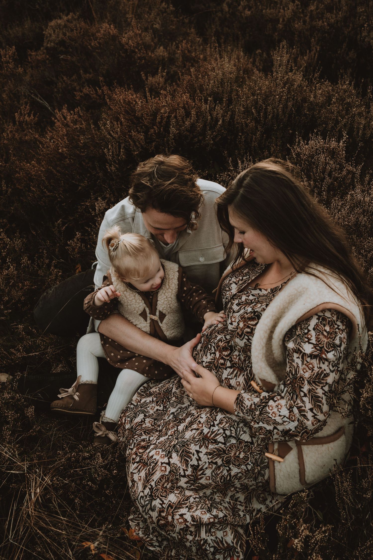 A family with a pregnant mother, father, and toddler sit together in a heathland field.