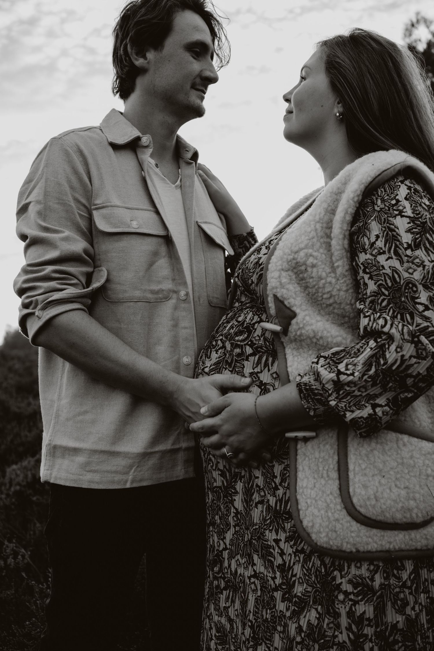 Black and white photo of a smiling couple holding hands, woman visibly pregnant, outdoors.