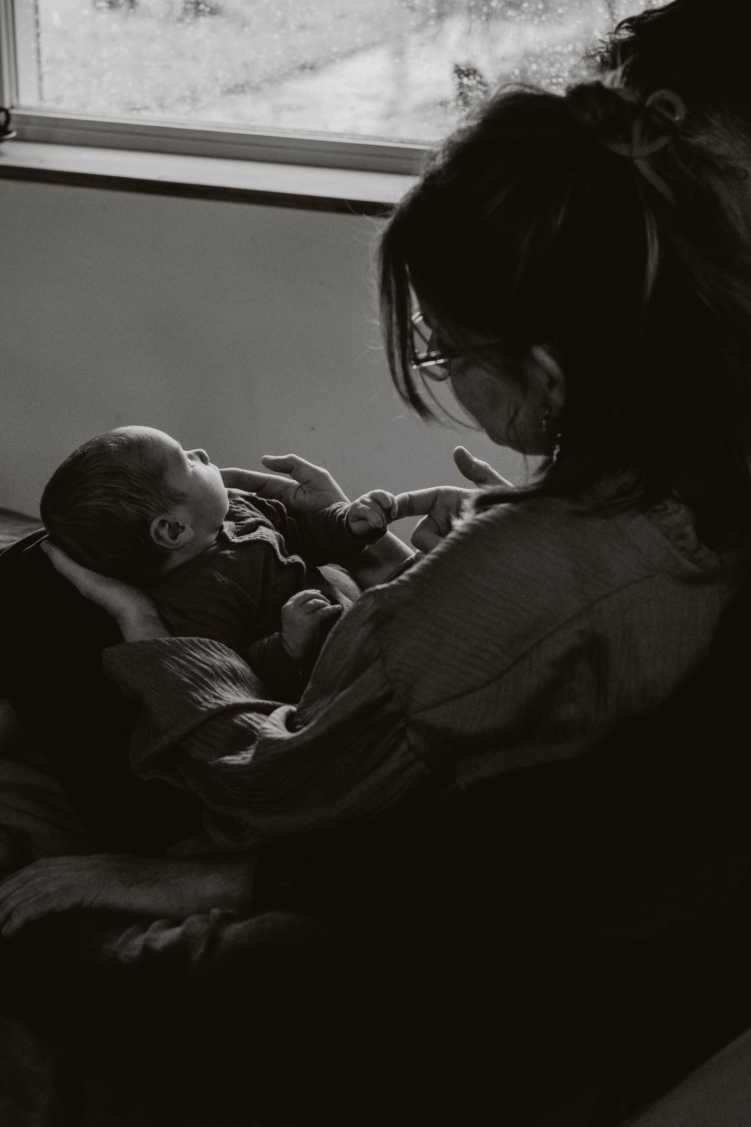 Black and white photo of mother holding newborn baby near a bright window.