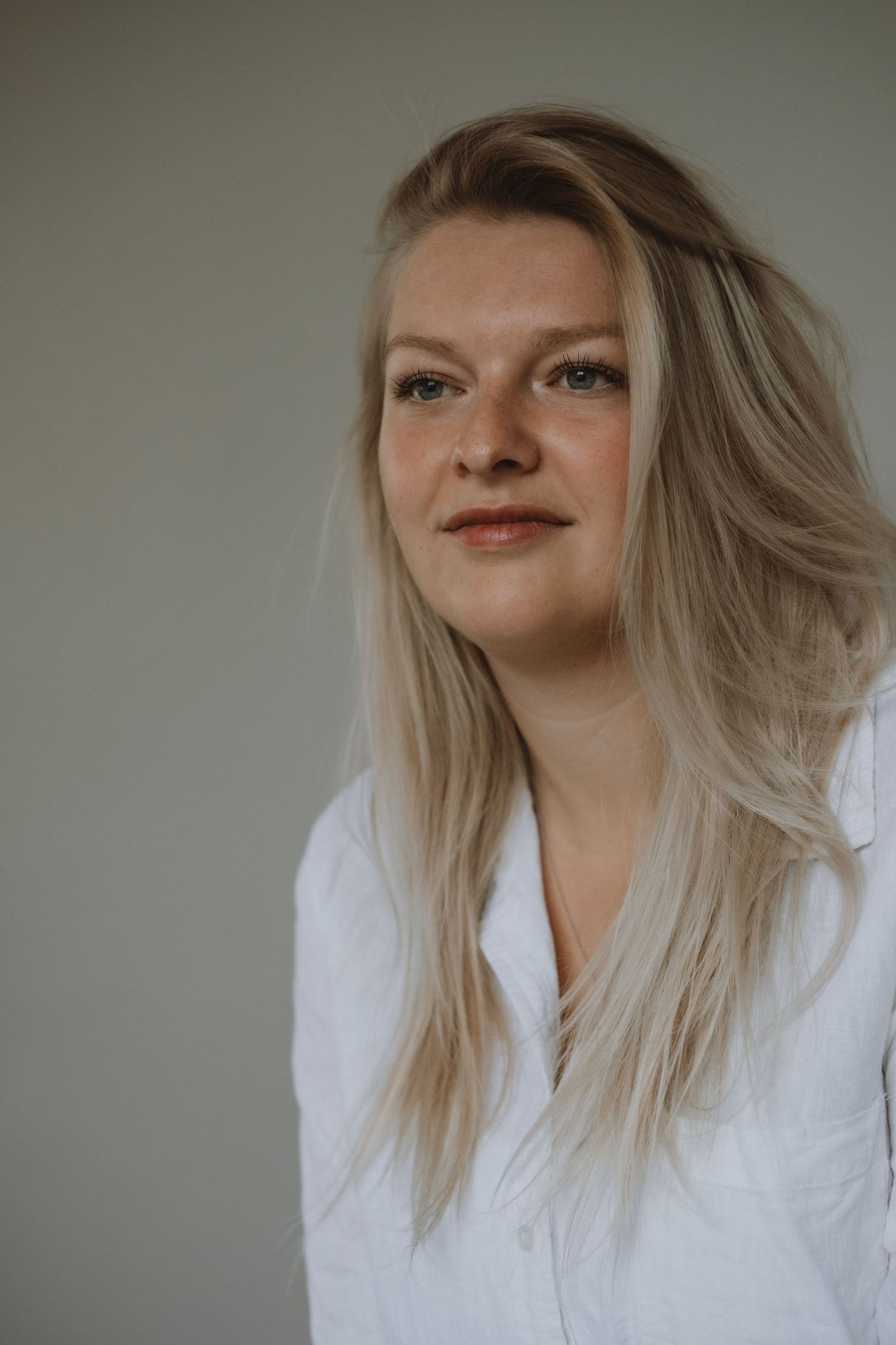 Blonde woman in white linen shirt with natural makeup poses against grey background.