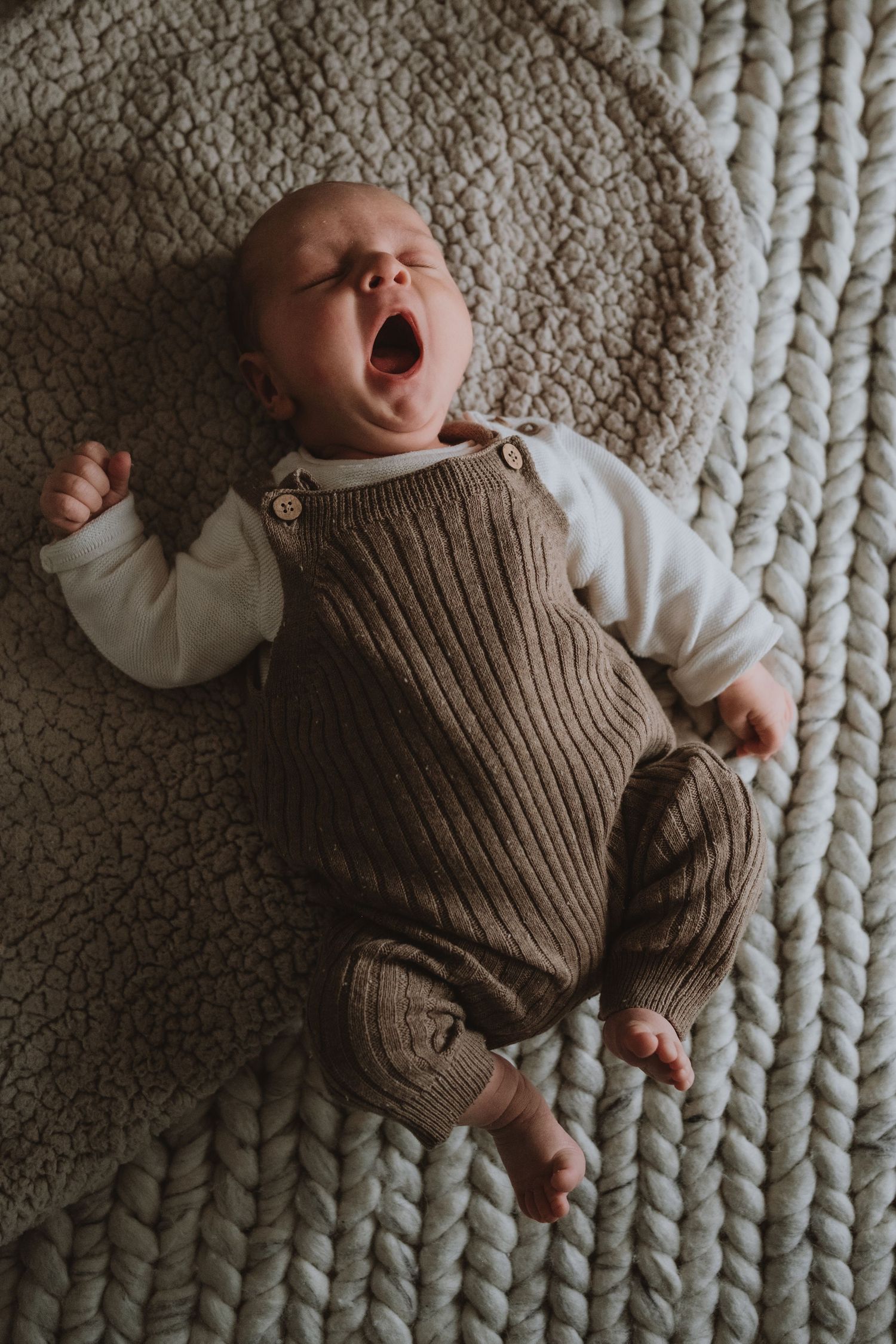 Adorable newborn baby yawning while lying on chunky knit blanket in brown knit overalls.