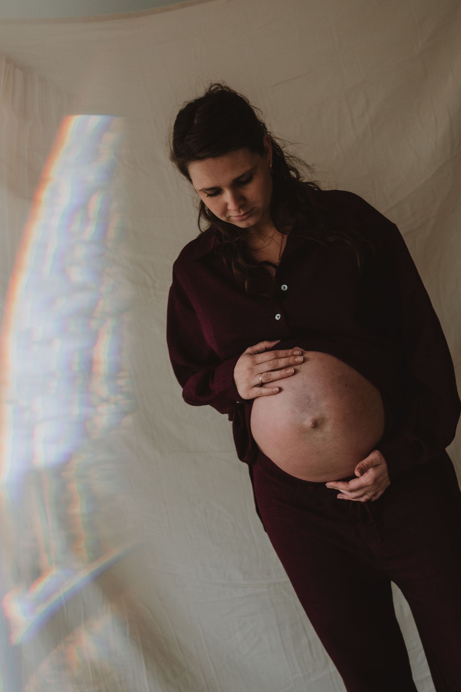 Pregnant woman in burgundy outfit holding bare baby bump with rainbow light effect.